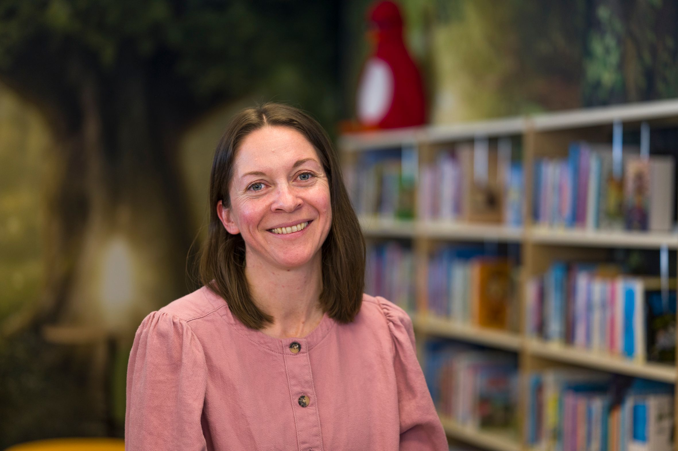 A smiling woman in a pink shirt stands in a library with bookshelves and a nature mural behind her.