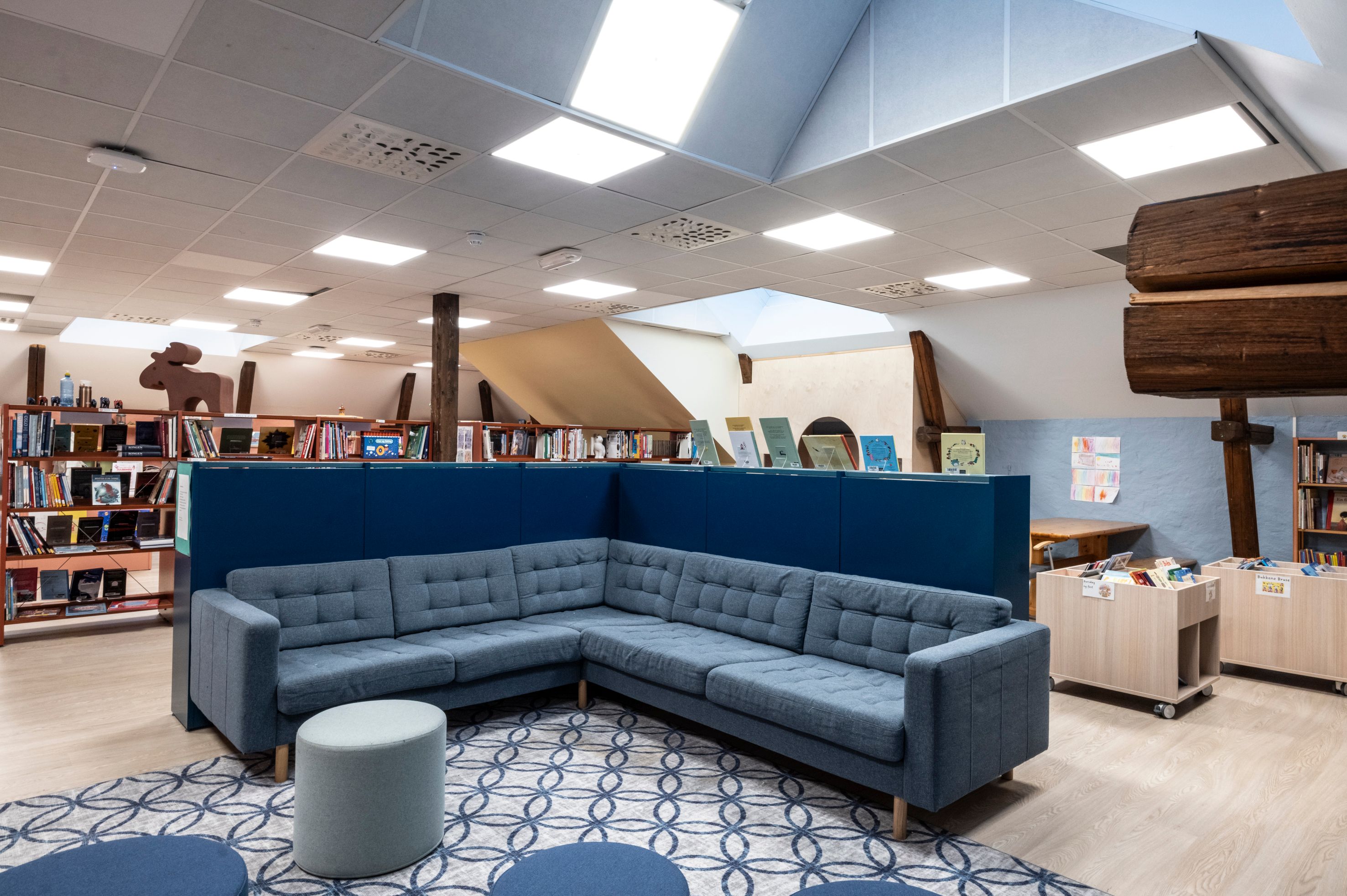 A modern, bright library or reading room with a blue L-shaped sofa, patterned rug, bookshelves, and visible wooden beams.