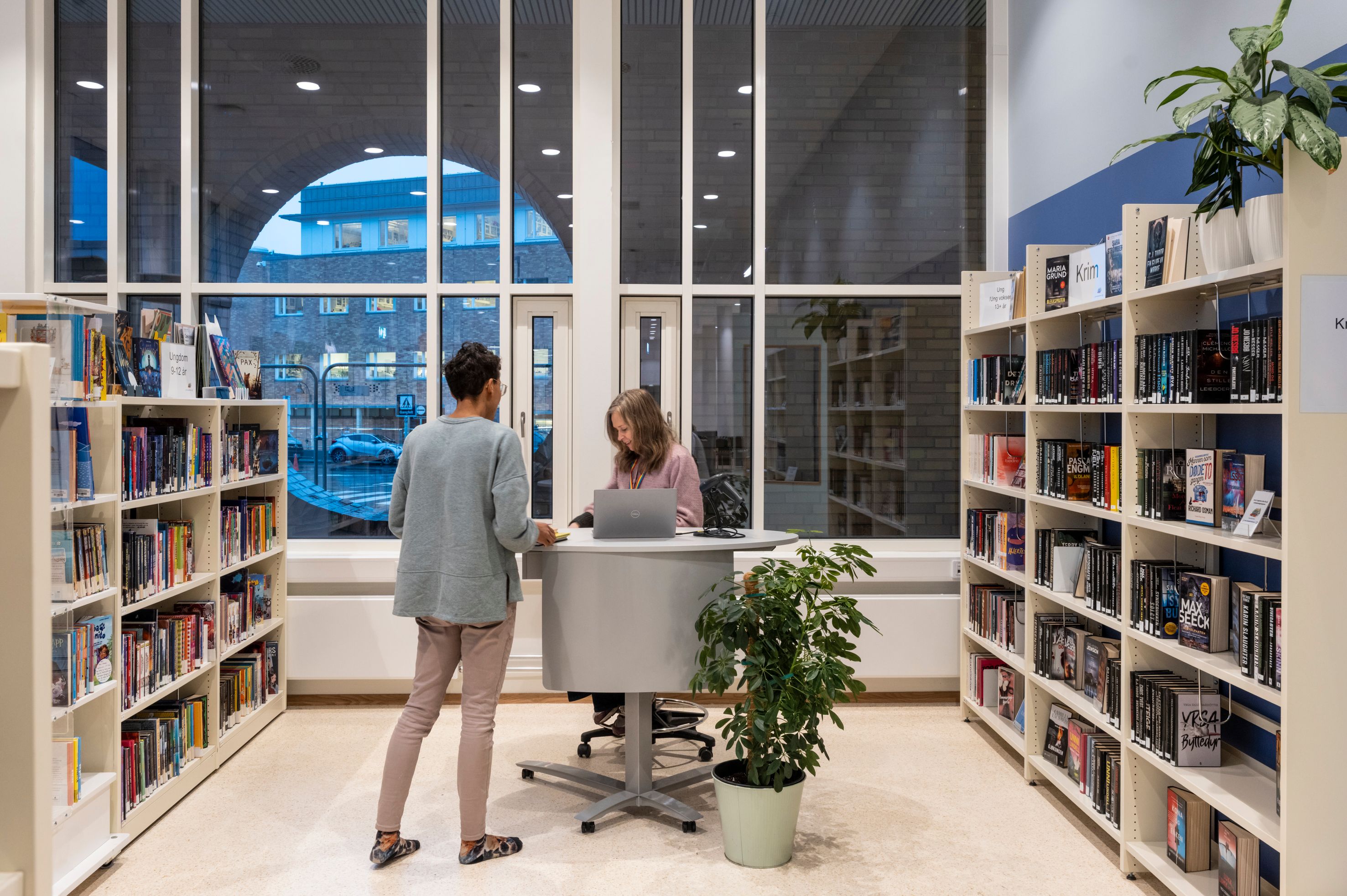 Two people at a service desk in a modern library, flanked by bookshelves and large windows.