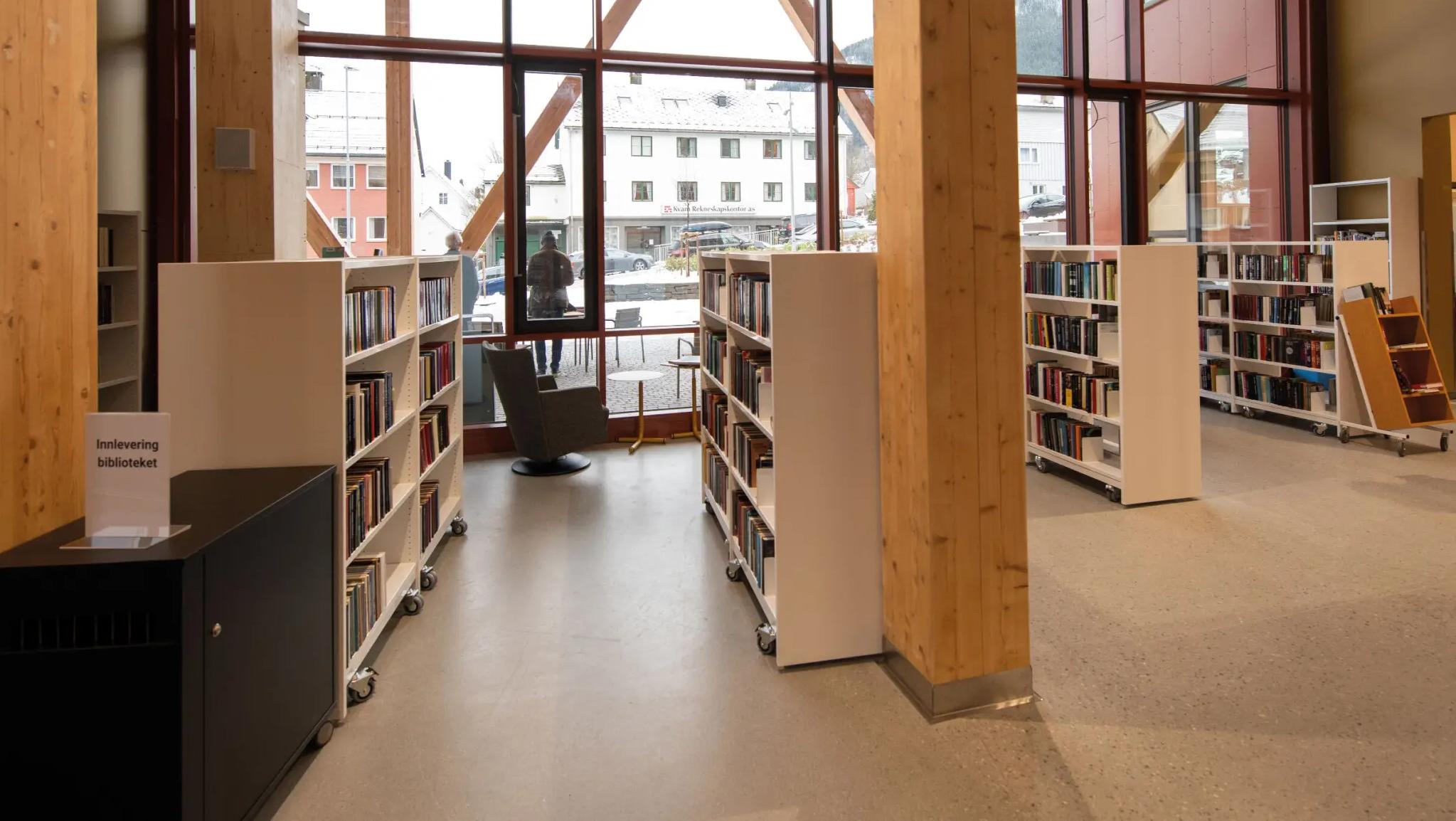 Modern library interior with large windows, wooden support beams, and white bookshelves filled with books.
