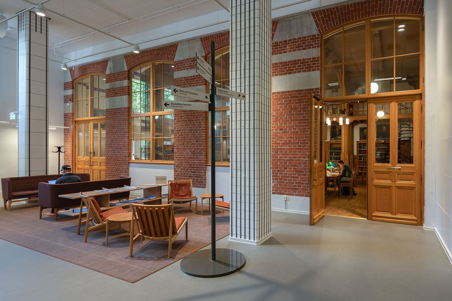 Spacious interior of a public building featuring a lounge area, red brick walls, large arched windows, and a person studying in an adjoining room through open wooden doors.