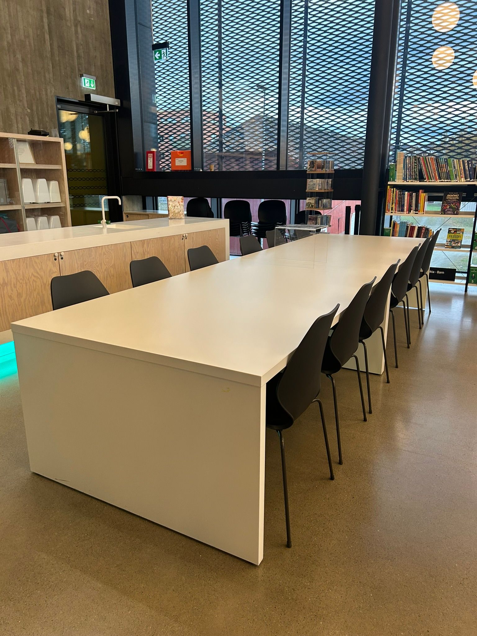 A long white table with black chairs in a bright, modern library or study area featuring large grid windows, bookshelves, and a counter.