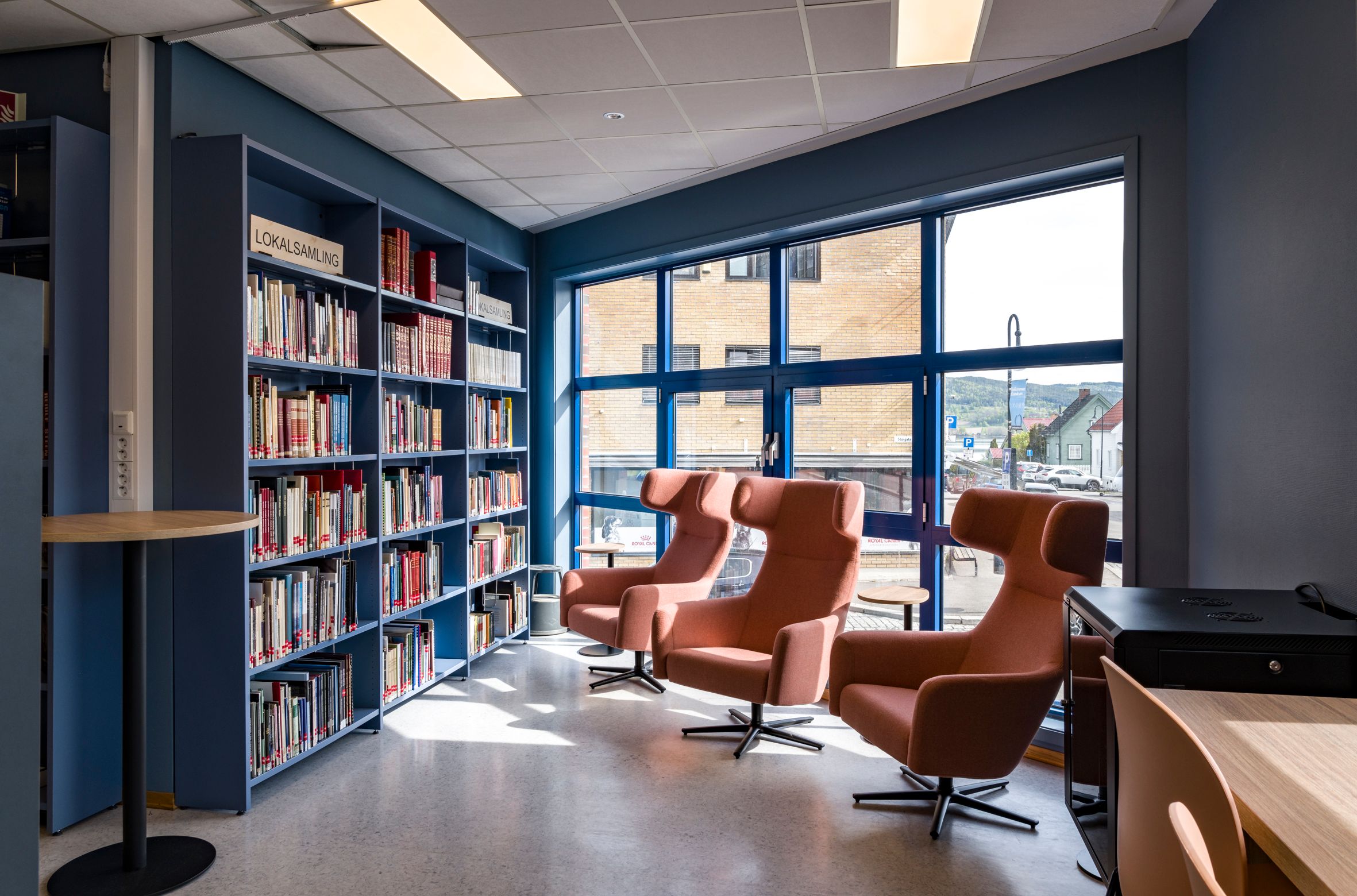 et bibliotek med en høy bokreoler langs veggen og 3 lenestoler foran de høye vinduene