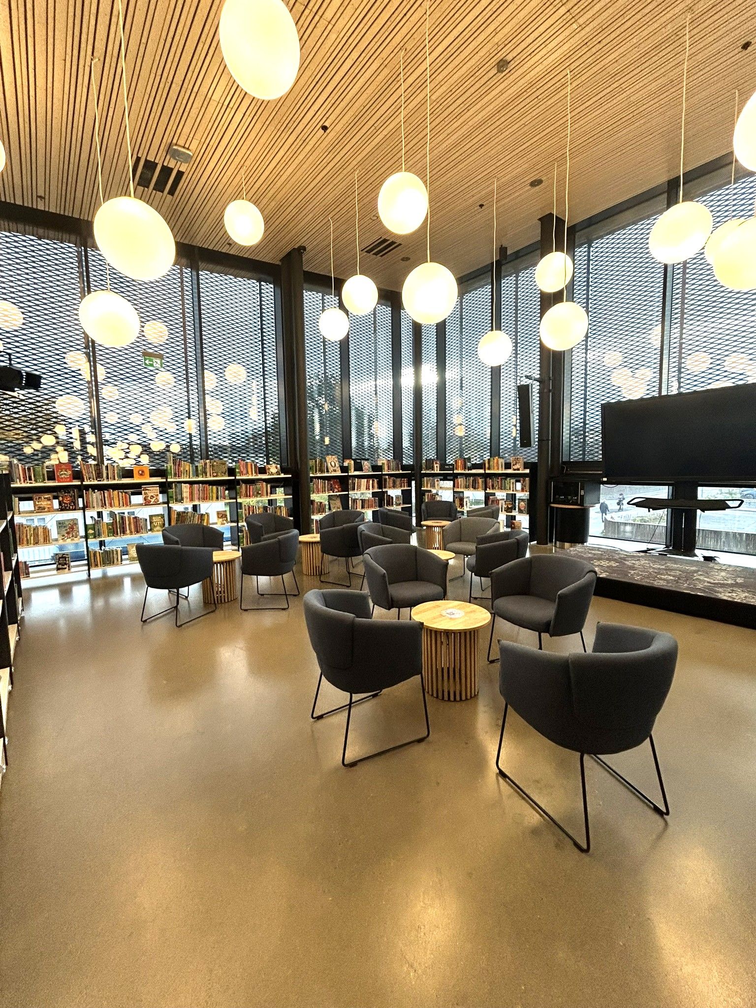Modern library lounge with dark armchairs, small wooden tables, bookshelves, large windows, and spherical pendant lights.