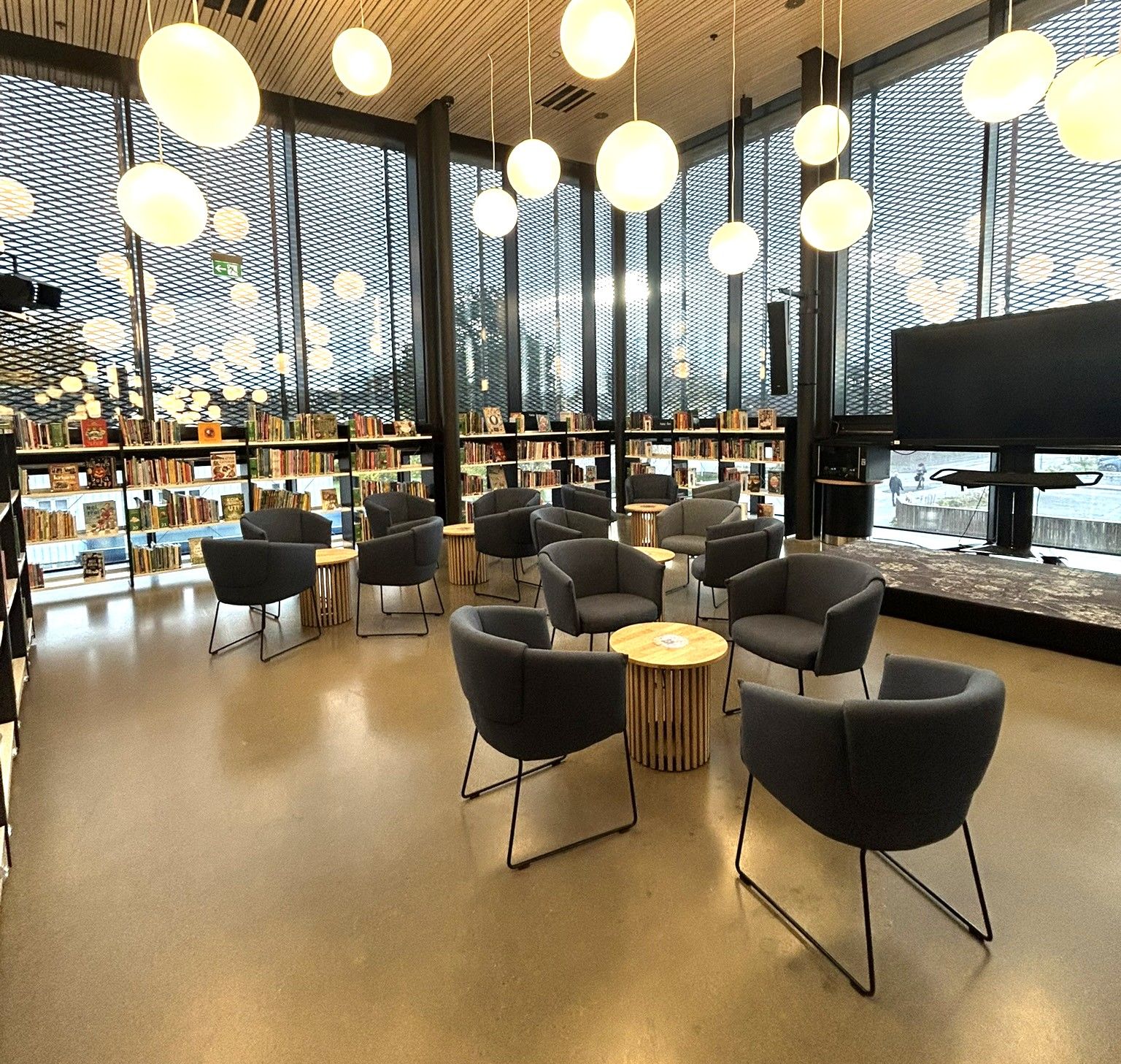 Modern library lounge with dark armchairs, small wooden tables, bookshelves, large windows, and spherical pendant lights.