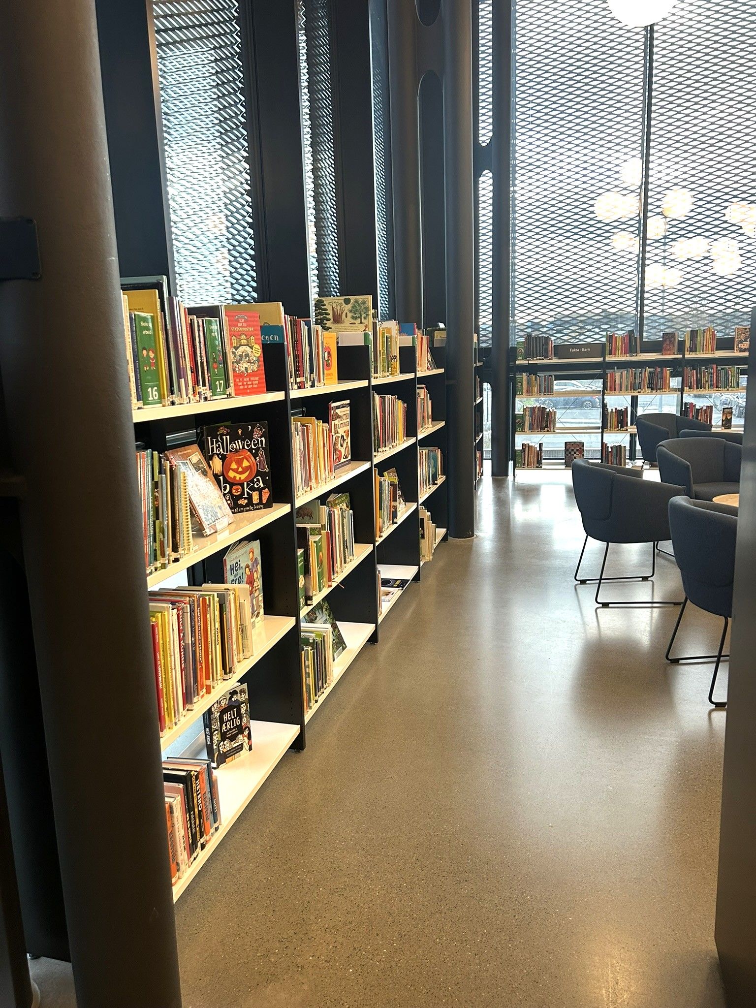 Modern library interior with tall bookshelves, a concrete floor, and a bright seating area by large patterned windows.