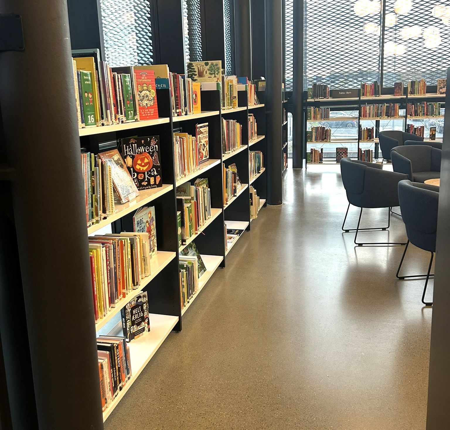Modern library interior with tall bookshelves, a concrete floor, and a bright seating area by large patterned windows.