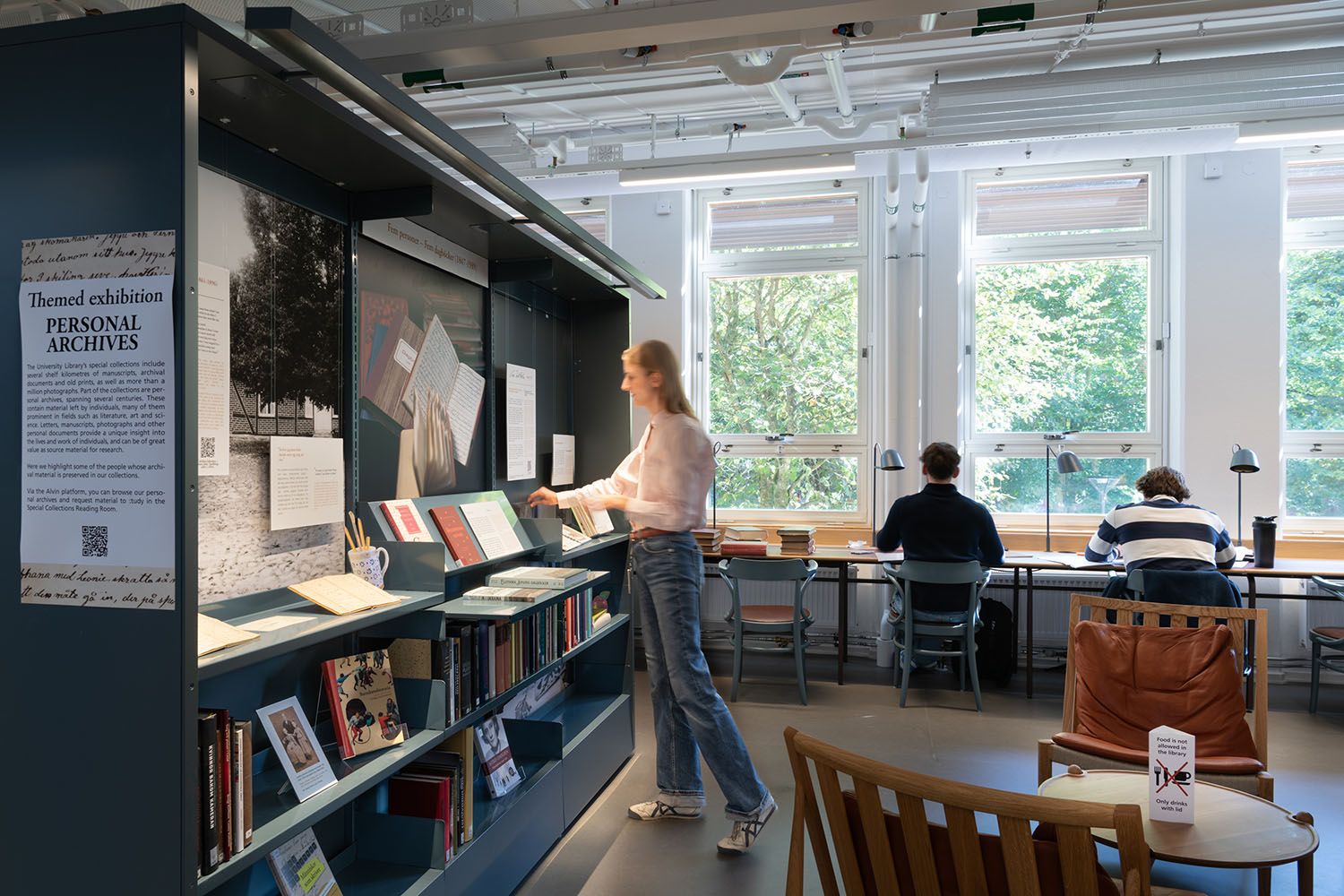 A woman browses a "Personal Archives" exhibition in a bright library with students studying at window desks.