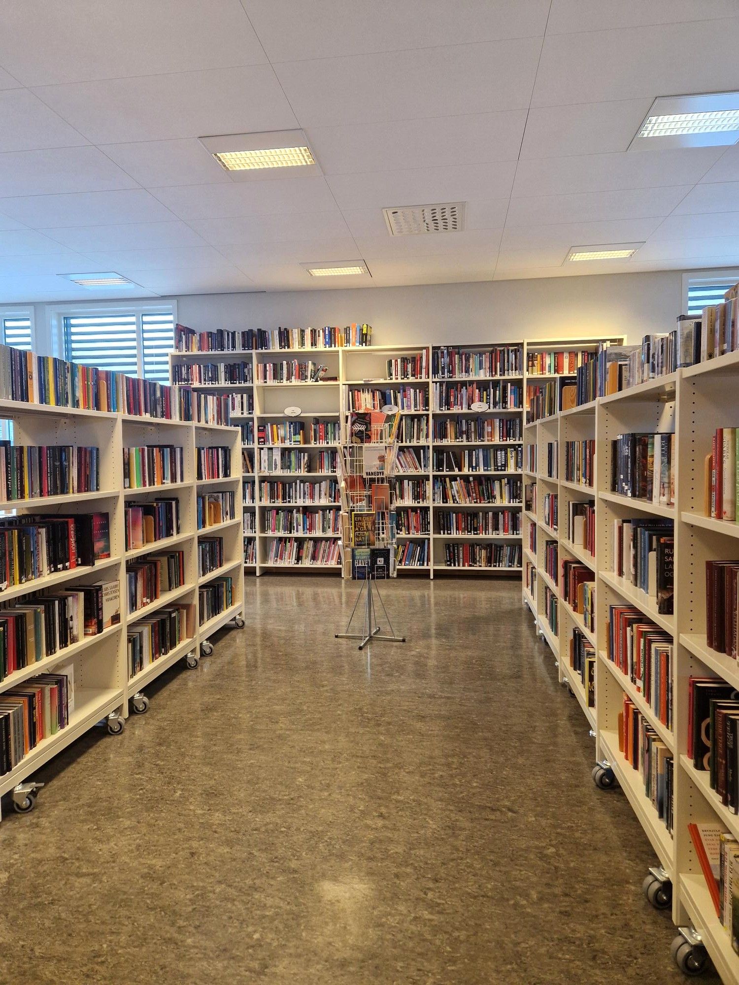 A brightly lit library aisle lined with tall white bookshelves packed with books, with a rotating display stand in the center.