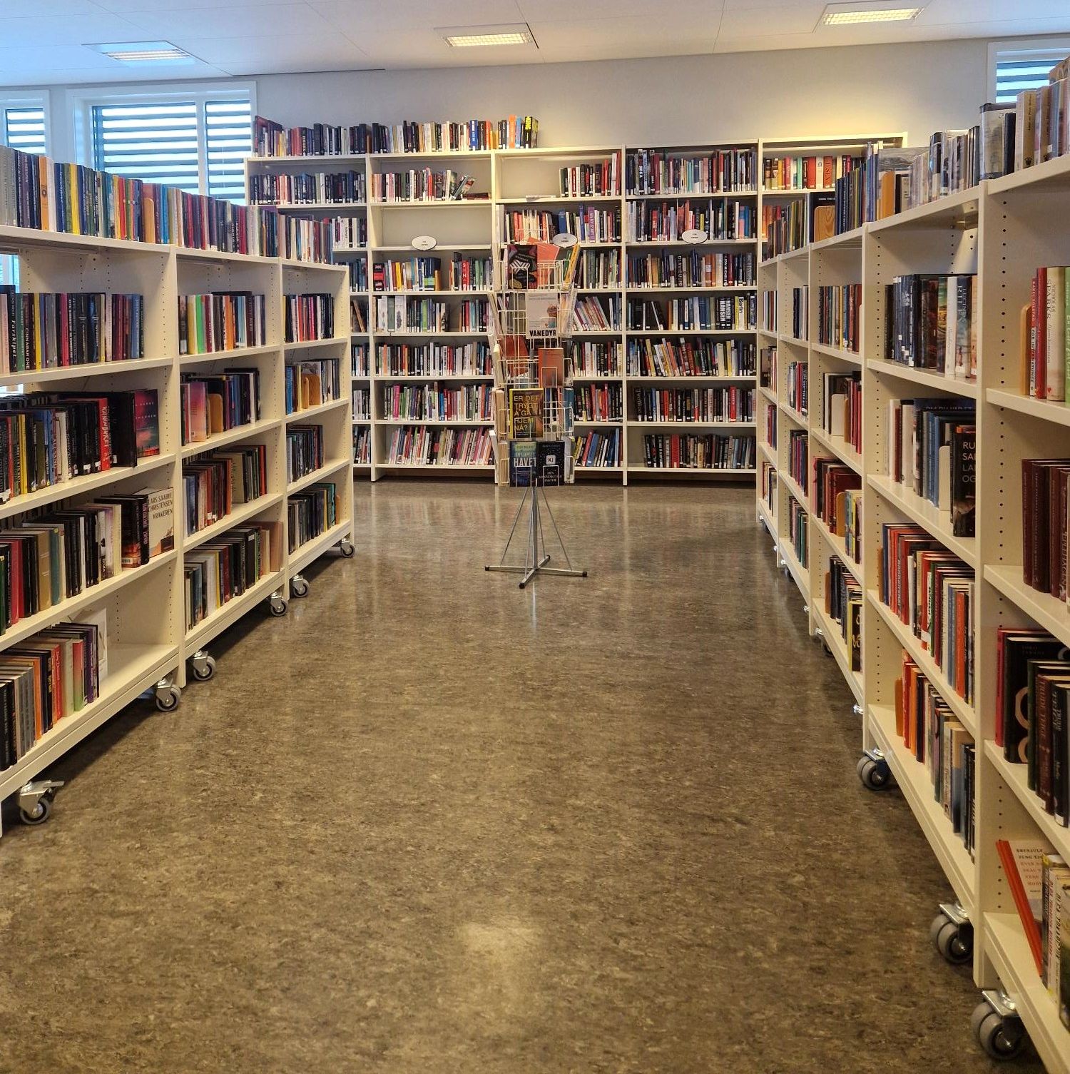 A brightly lit library aisle lined with tall white bookshelves packed with books, with a rotating display stand in the center.