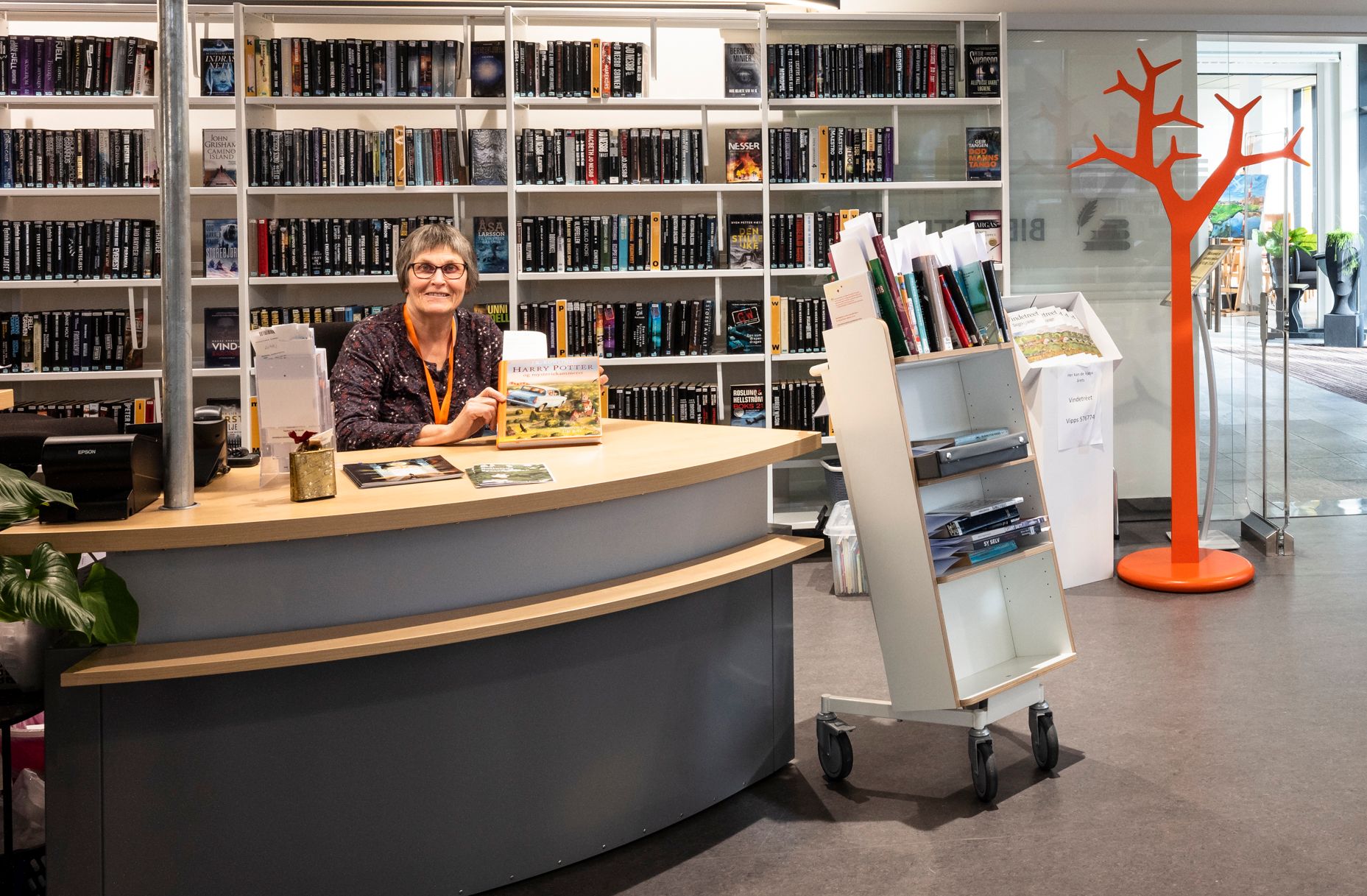 en dame sitter ved en grå bibliotekskranke med en bokvogn foran