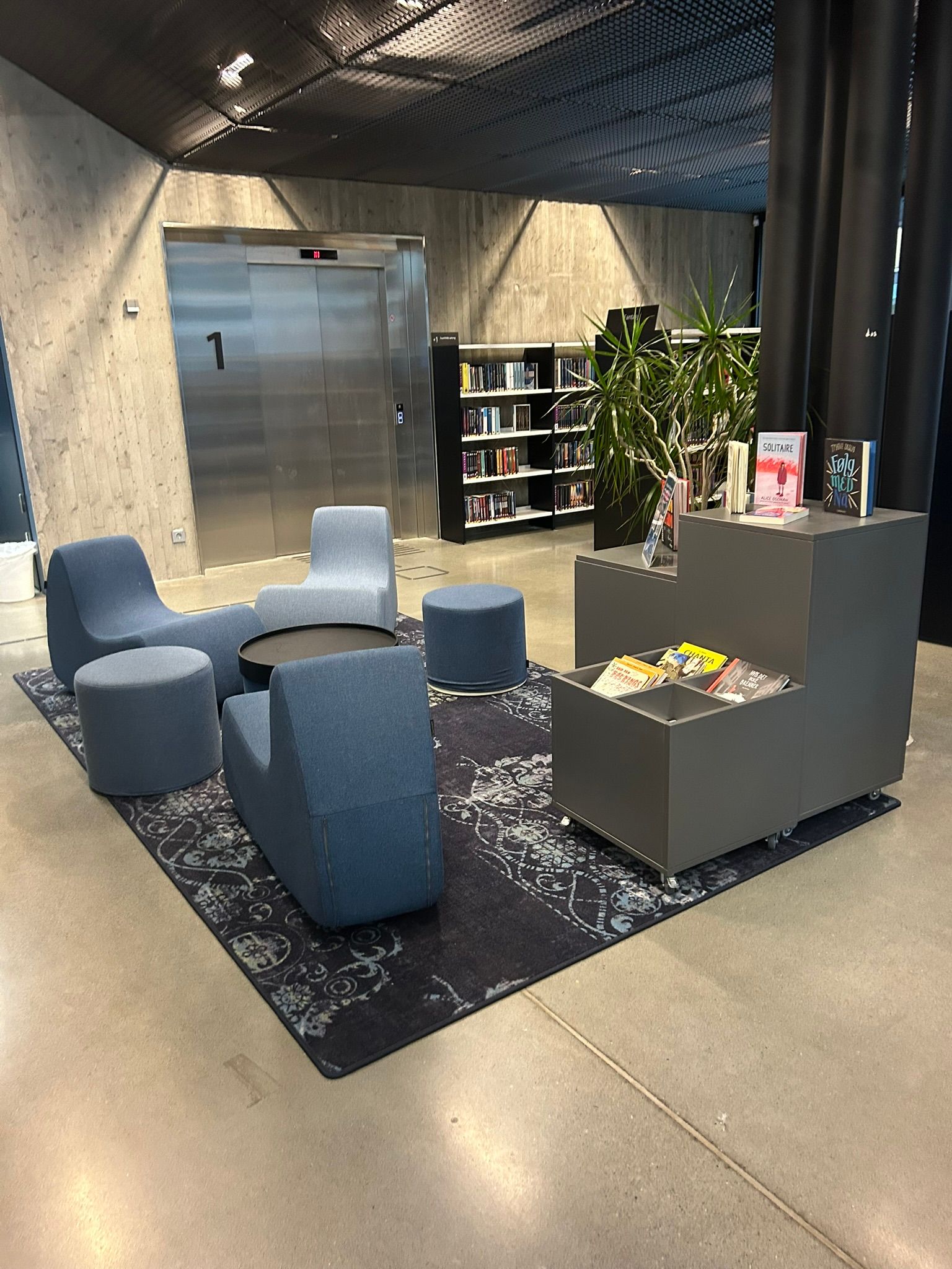 A modern library lounge with blue modular seating, a bookshelf, and a large metal elevator.