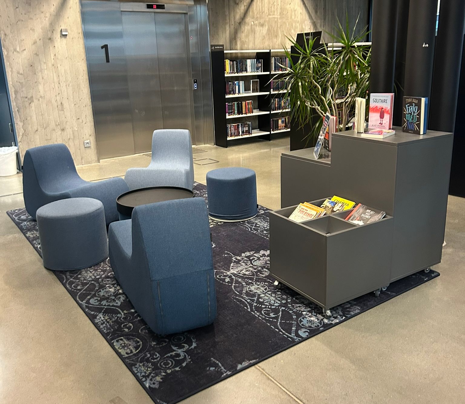 A modern lounge area with blue modular seating on a patterned rug, a gray book display, an elevator, and bookshelves.