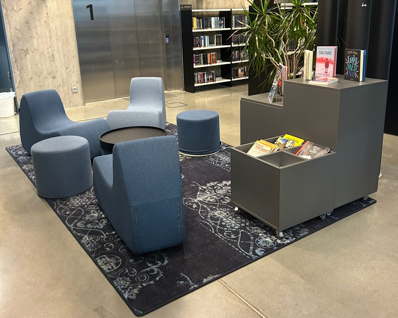 A modern library lounge with blue modular seating, a bookshelf, and a large metal elevator.