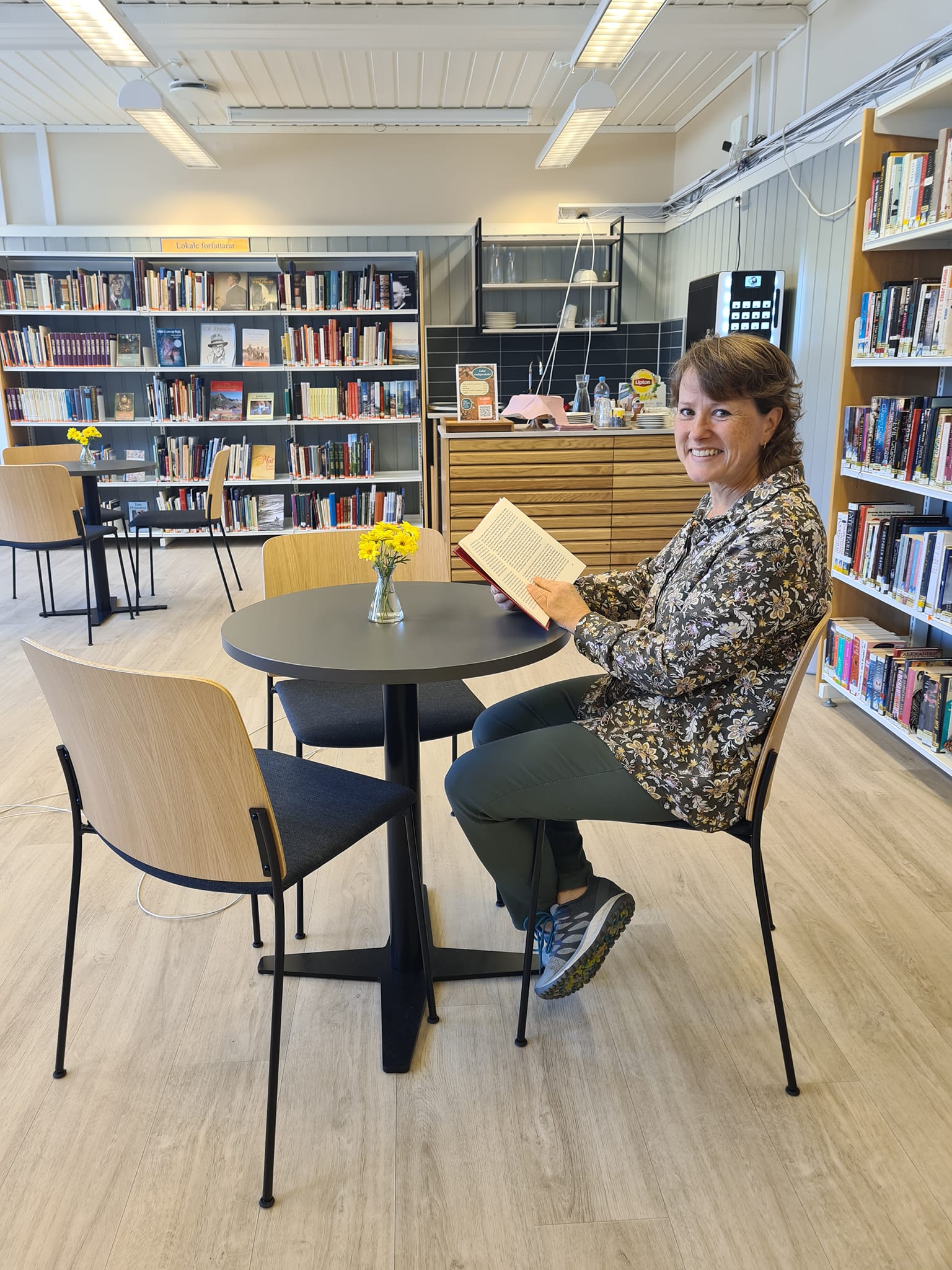 A smiling woman sits at a table in a library, holding an open book.