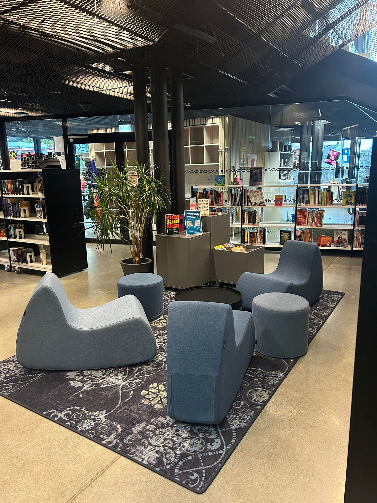 Modern library interior featuring a seating area with blue lounge chairs and ottomans on a patterned rug, surrounded by bookshelves.