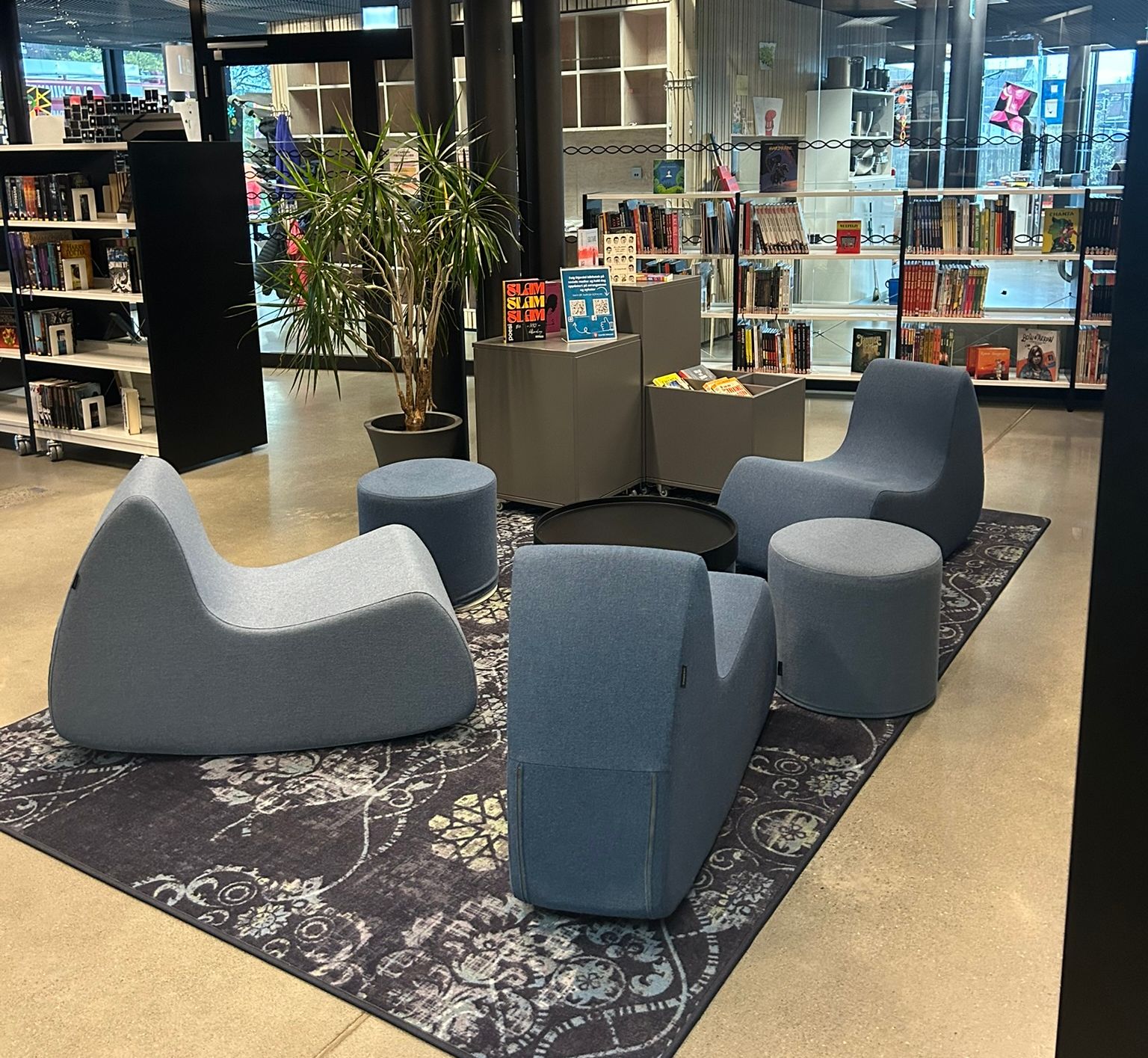 Modern library interior featuring a seating area with blue lounge chairs and ottomans on a patterned rug, surrounded by bookshelves.