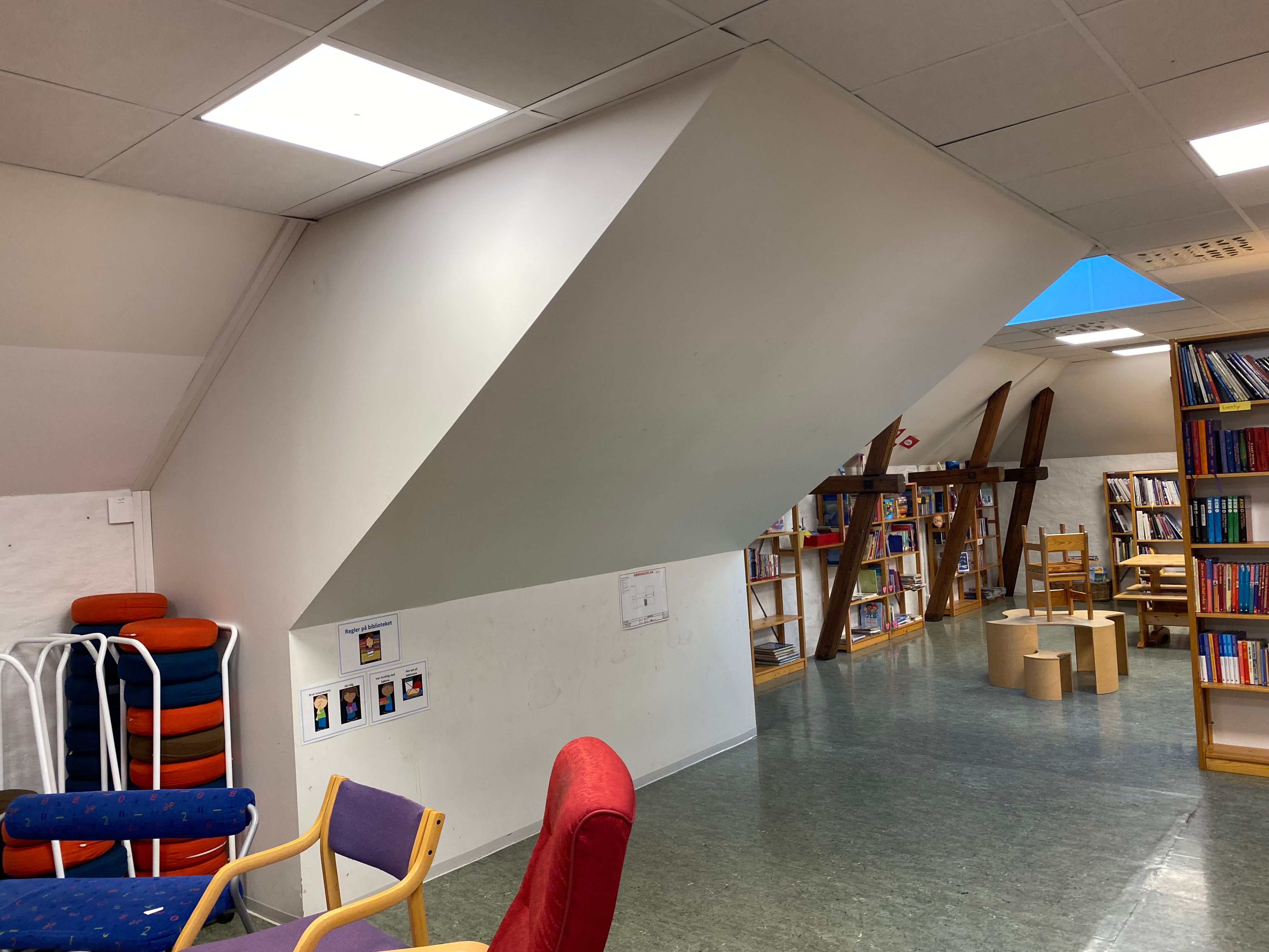An attic-style library with bookshelves, various chairs, and a skylight revealing blue sky.