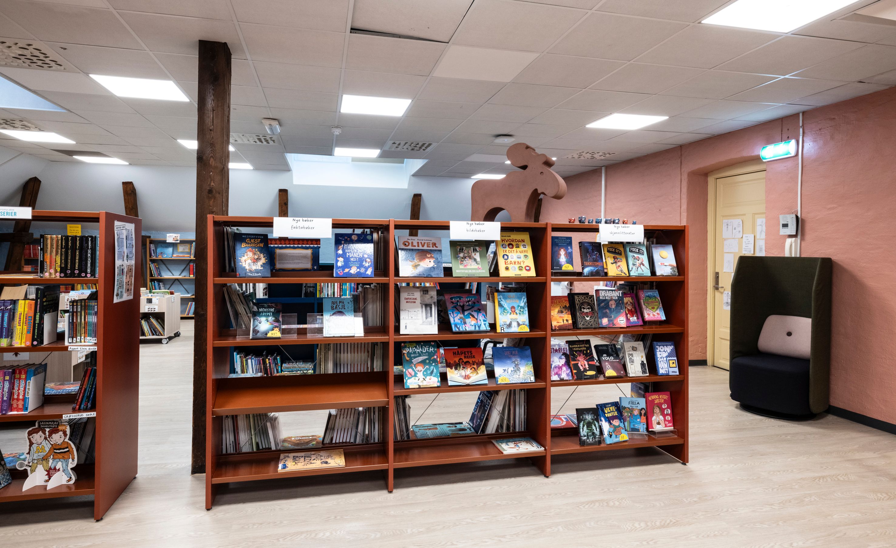 A children's library section with wooden bookshelves full of colorful books, a pink wall, a dark green reading chair, and a moose cutout.
