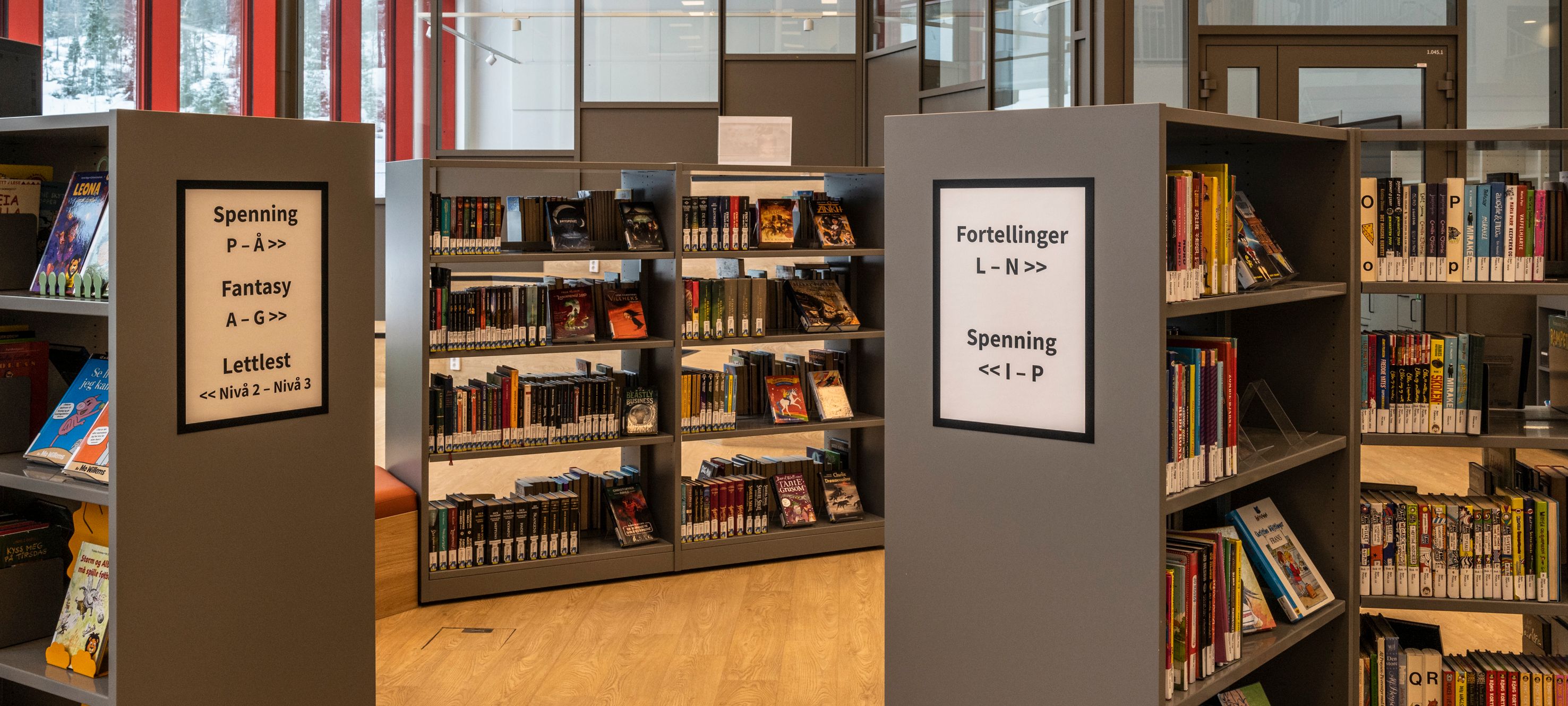 A modern library with grey bookshelves filled with books, organized by categories like "Spennning" (thriller) and "Fantasy" in Norwegian.