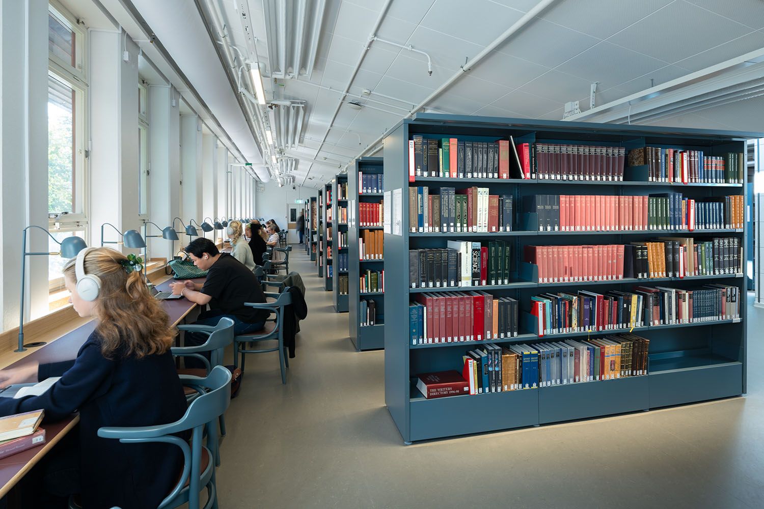 A modern library with people studying at desks by large windows, and rows of bookshelves on the right.