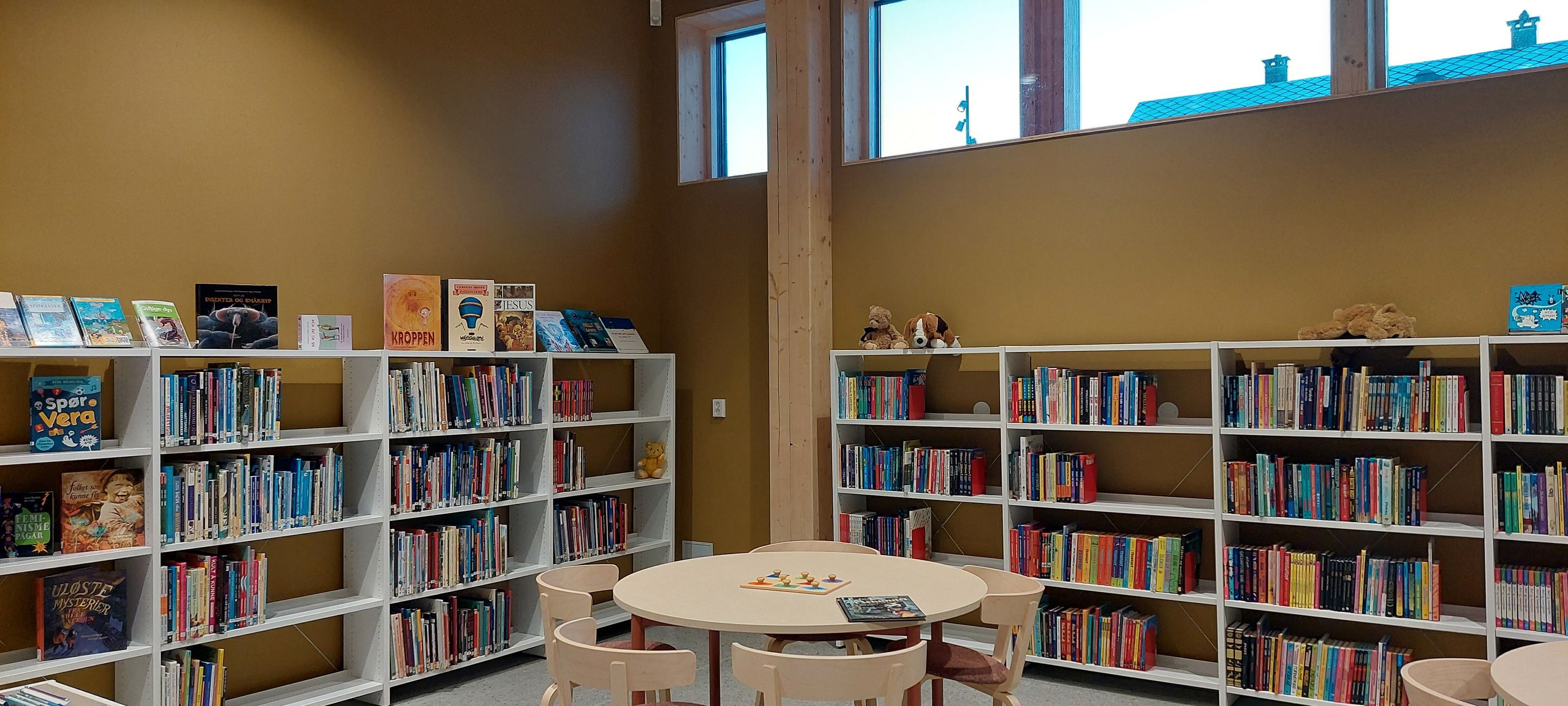 Children's library with white bookshelves full of books, a round table with small chairs, and high windows.