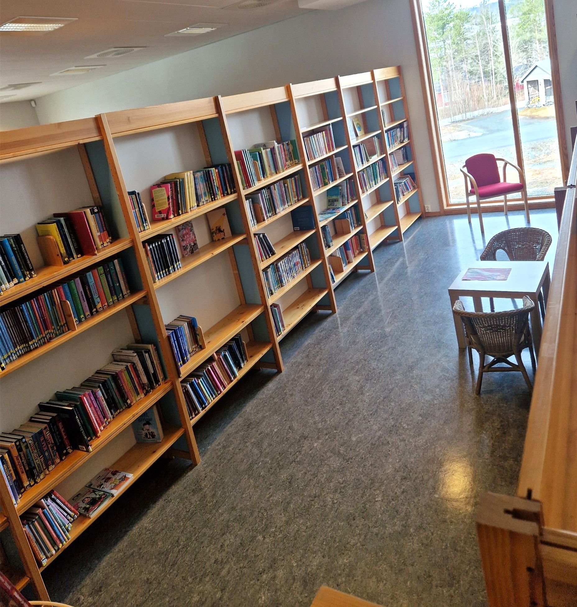 A library room with bookshelves, a large window looking out to trees, and a table with wicker chairs.
