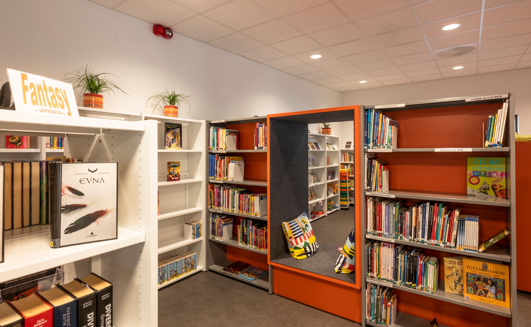 A modern library space with white and orange bookshelves, a reading nook, and a "Fantasy" section sign.