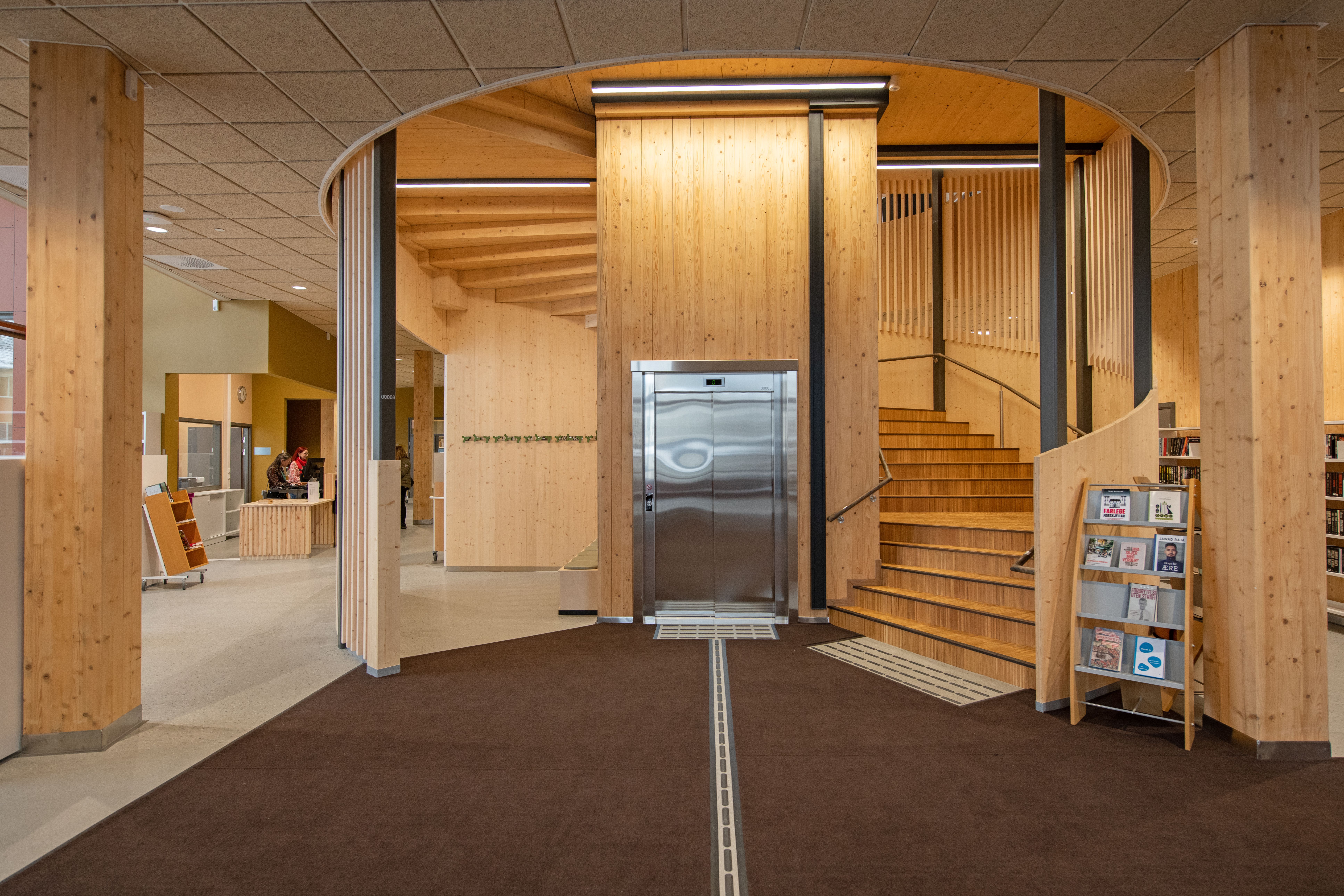 Modern interior with light wood paneling, a curved staircase and elevator, and a reception counter.