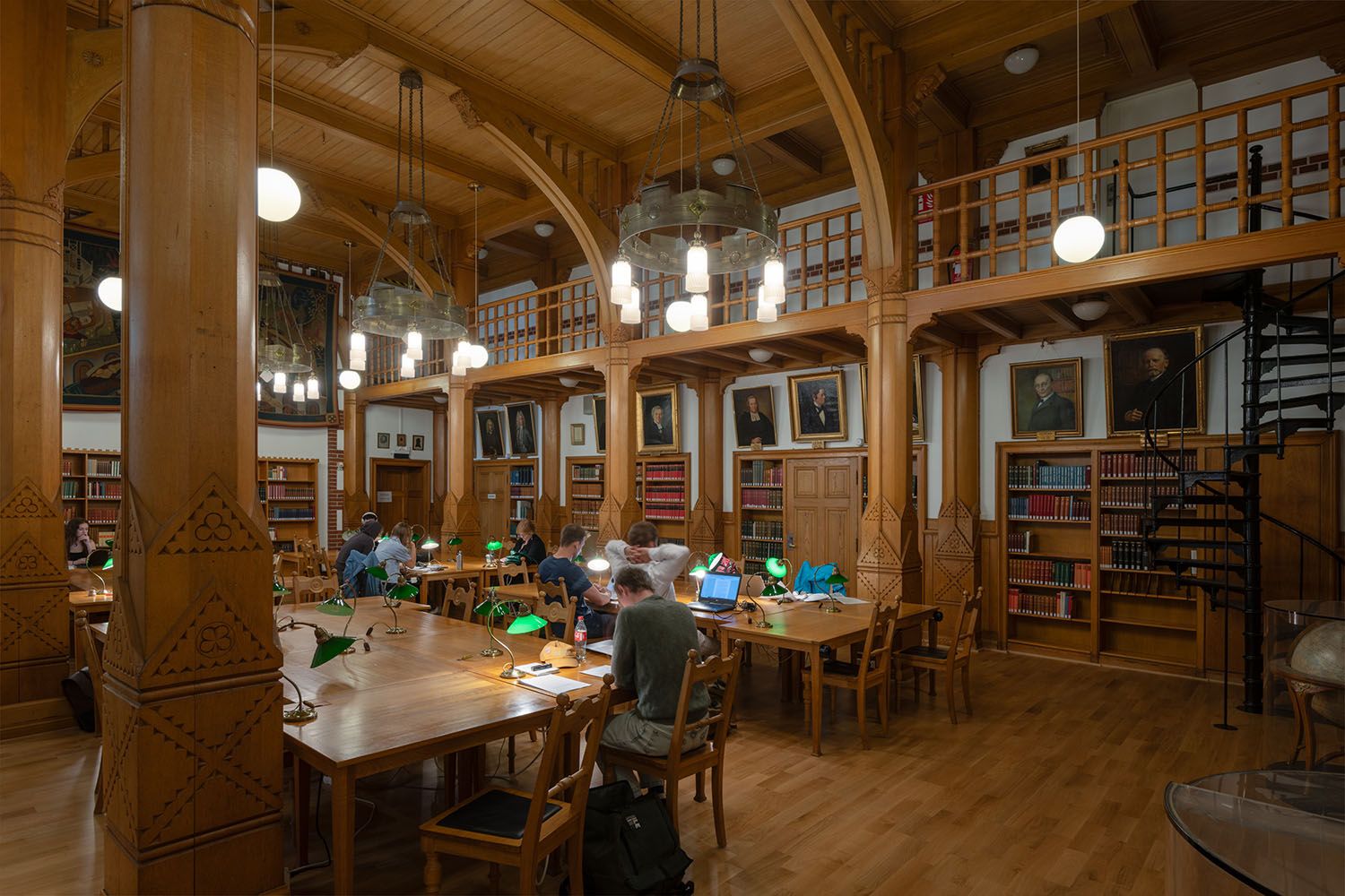 A spacious, wood-paneled library with students studying at tables, surrounded by bookshelves, portraits, and a spiral staircase.