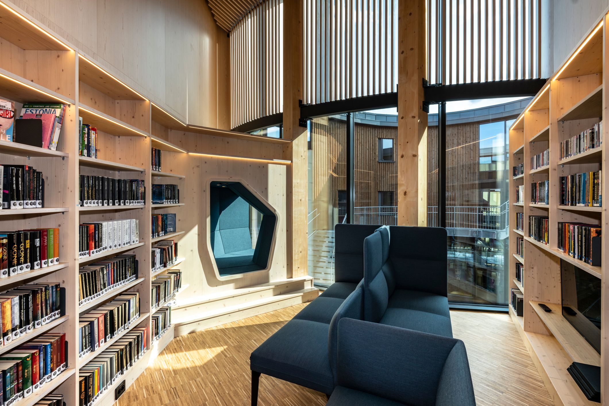 A modern reading room with light wooden bookshelves, a blue modular sofa, a built-in reading nook, and large windows overlooking other wooden buildings.