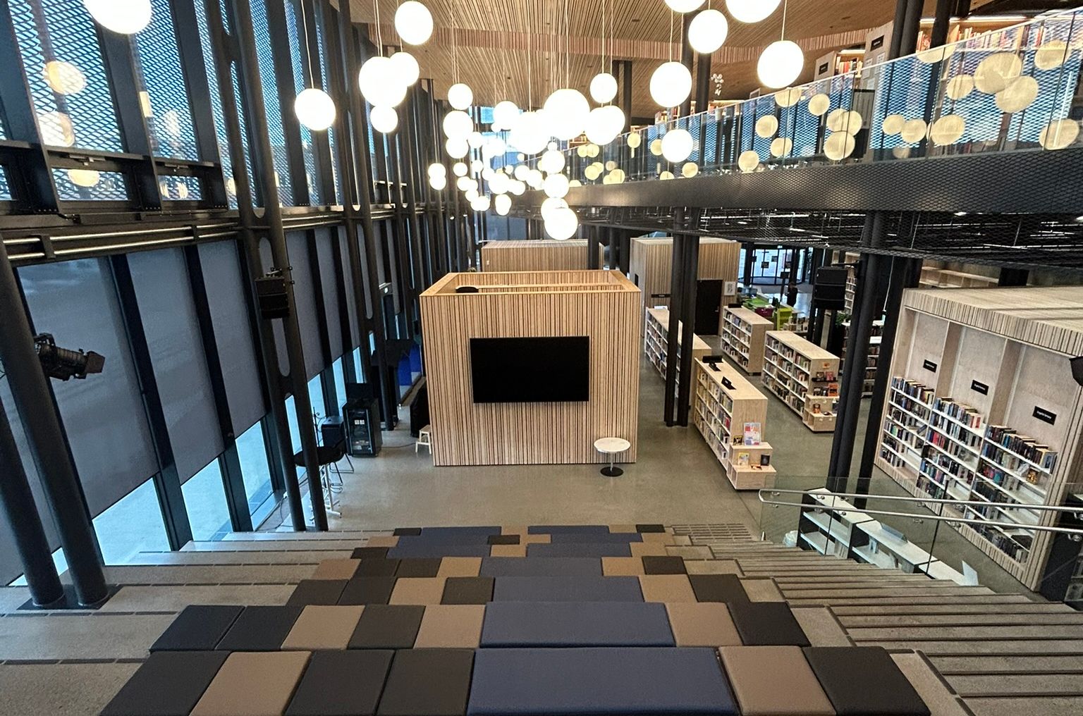 Modern library interior with tiered seating, a wooden slatted ceiling with numerous globe lights, and bookshelves on multiple levels.