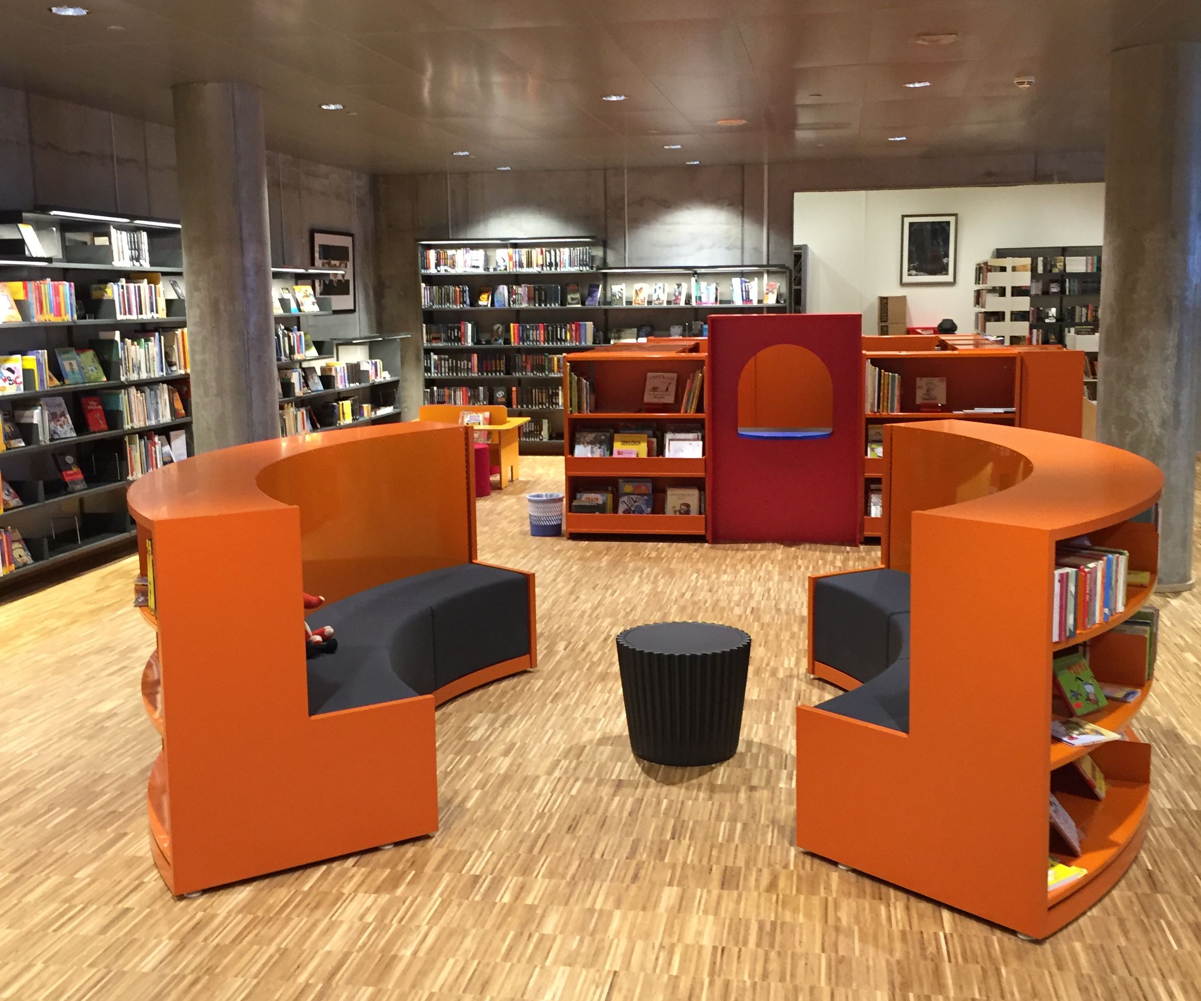 et bibliotek med oransje stoler og bokhyller og et tregulv.