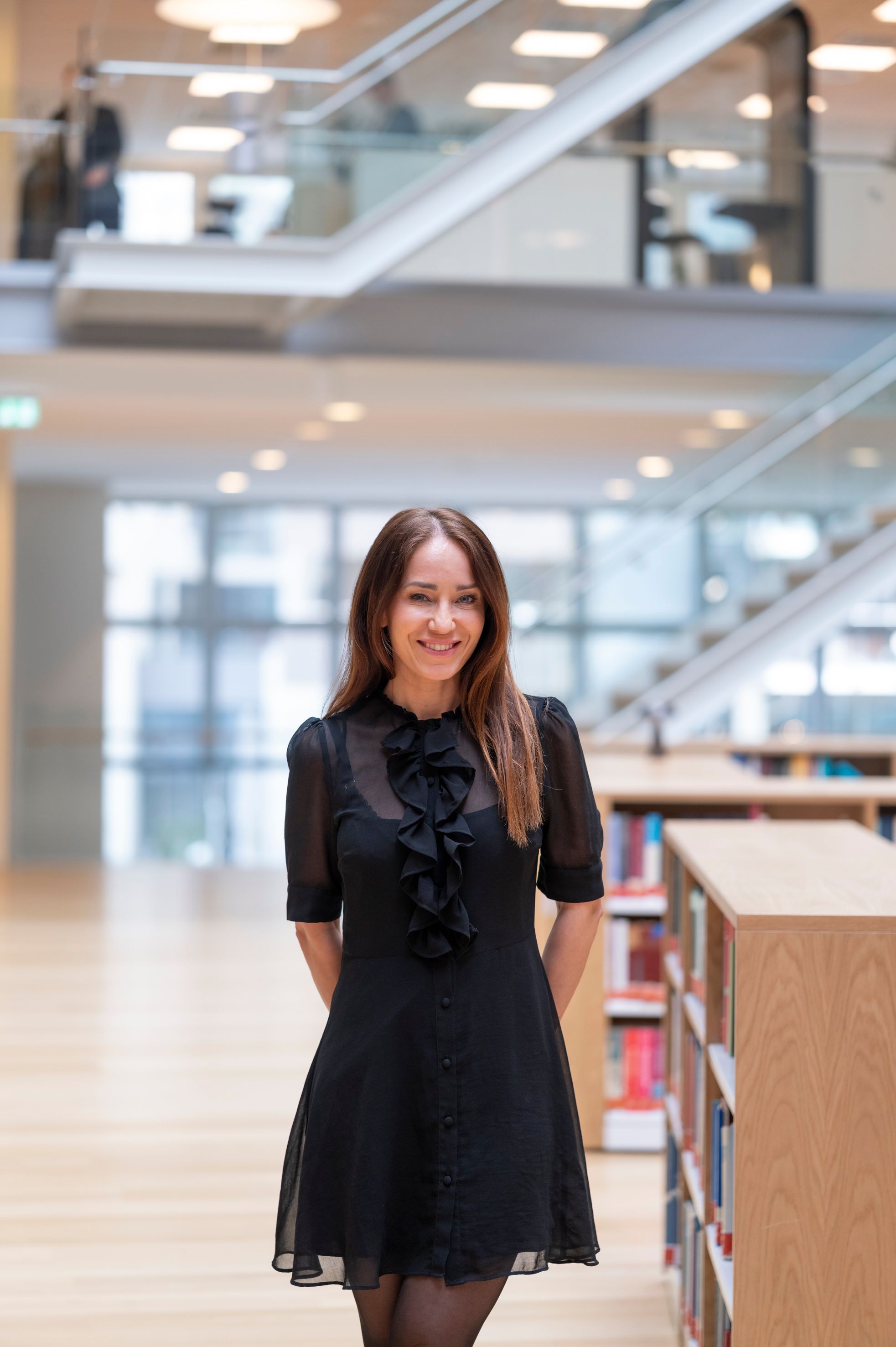 A smiling woman in a black ruffled dress stands in a modern library.
