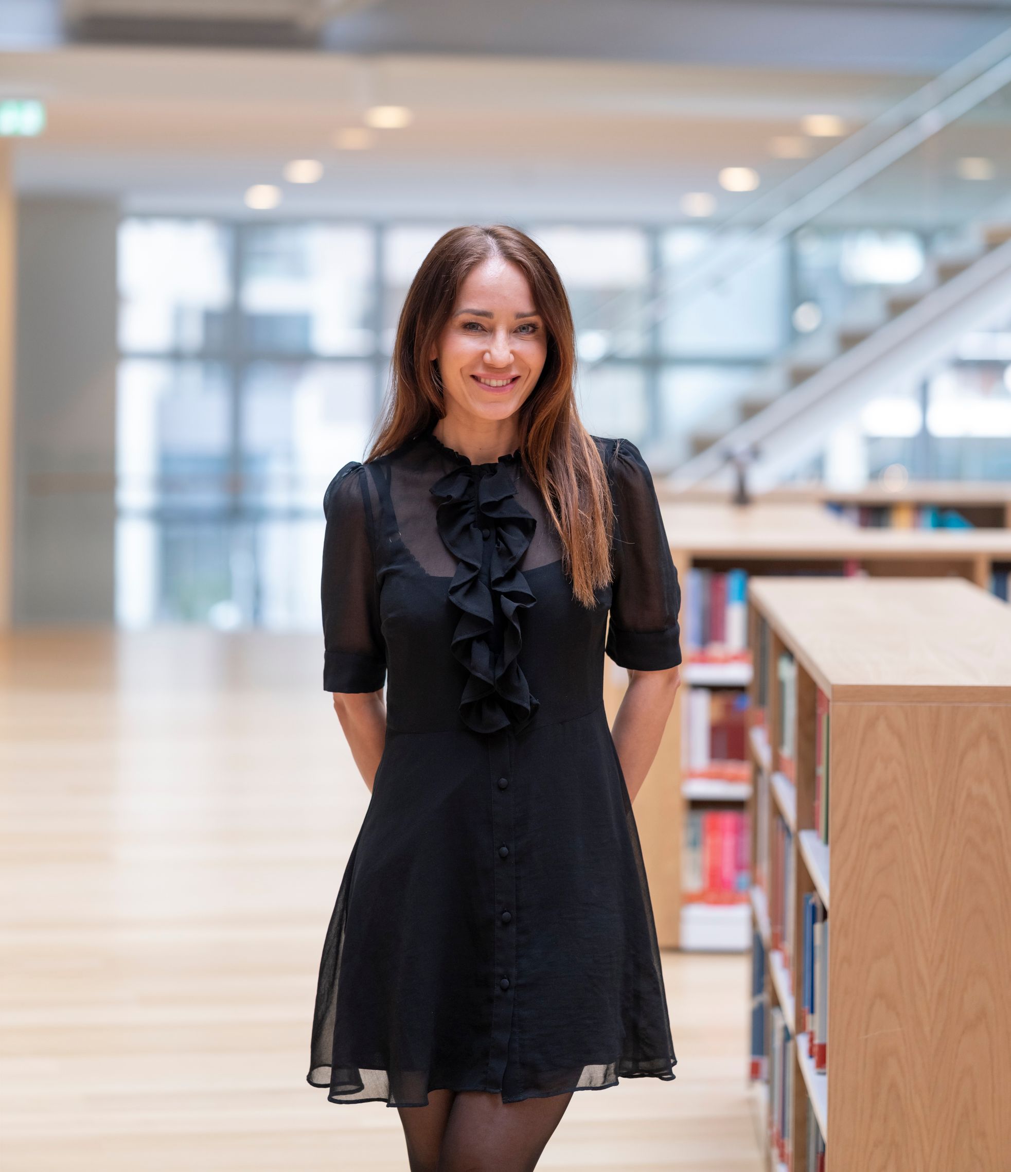A smiling woman in a black ruffled dress stands in a modern library.