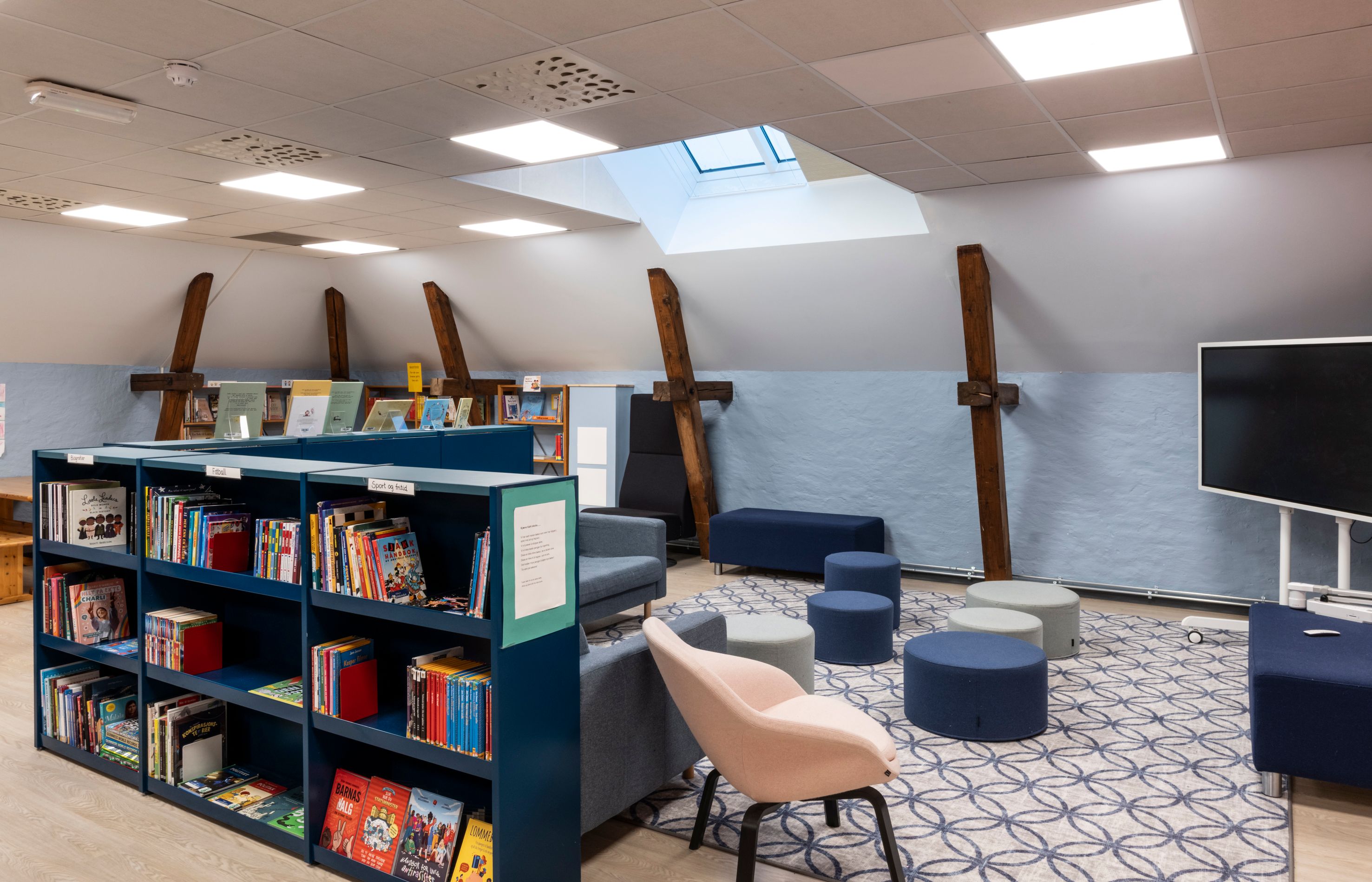 Modern attic library with blue bookshelves, varied seating, exposed wooden beams, skylights, and a large TV.