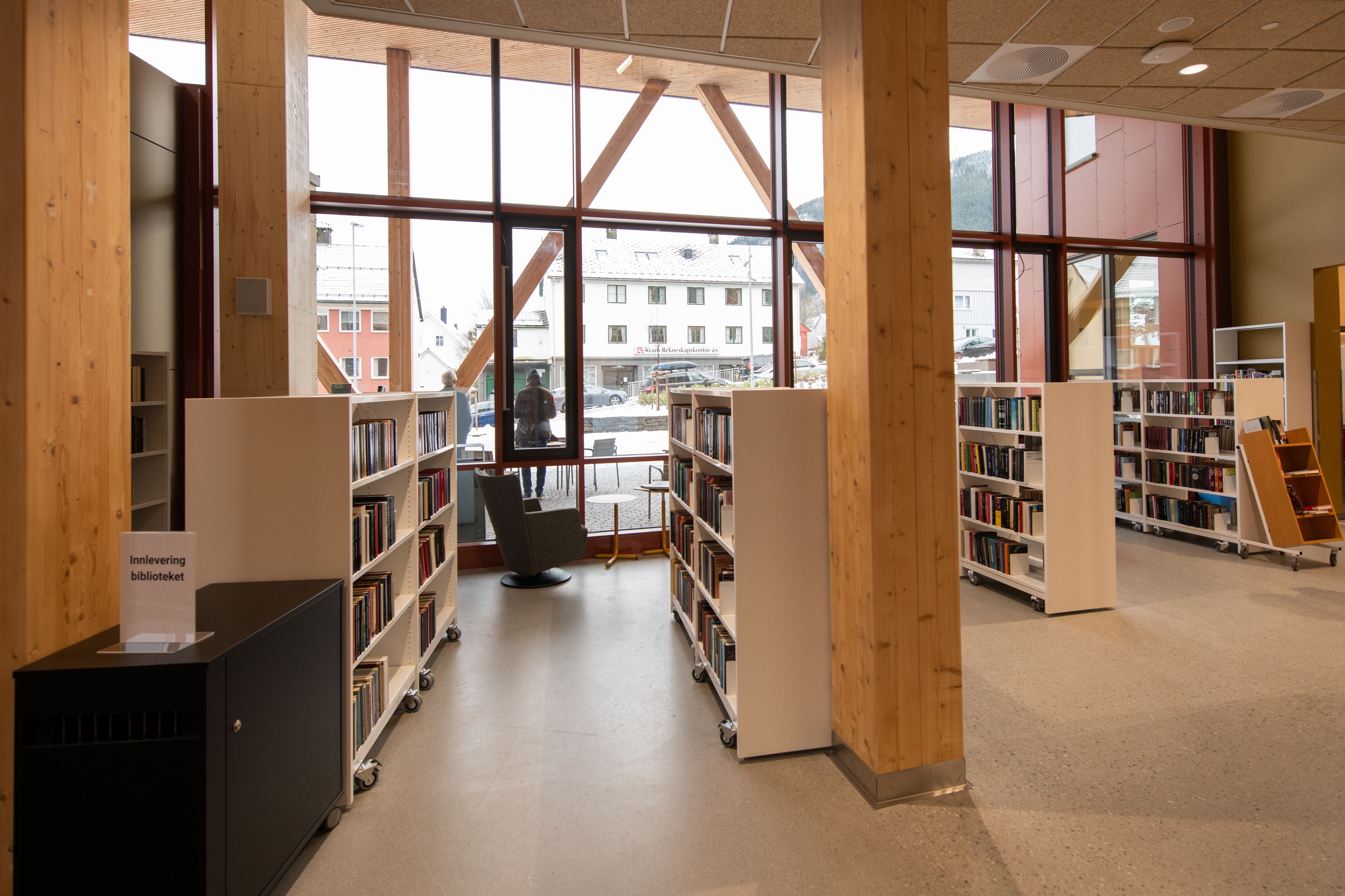 A modern library interior with wooden pillars, mobile bookshelves, and large windows looking out onto buildings.