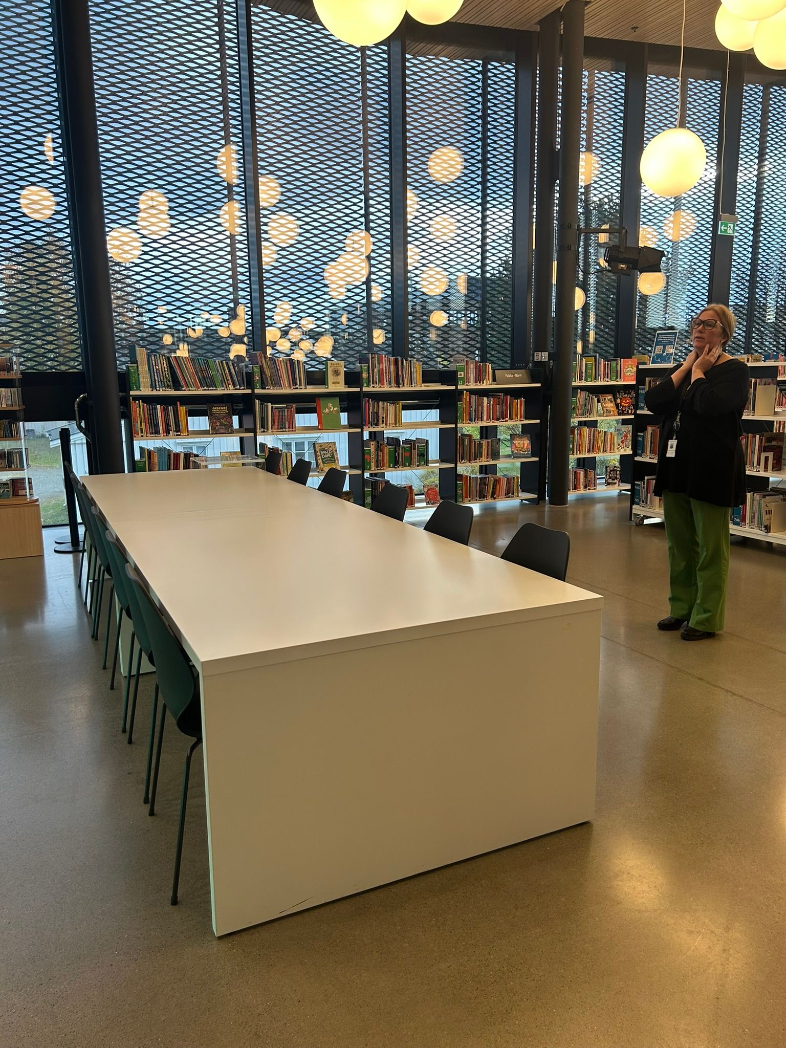 A woman stands in a modern library with a long white table, bookshelves, and large windows with a mesh pattern, under spherical ceiling lights.