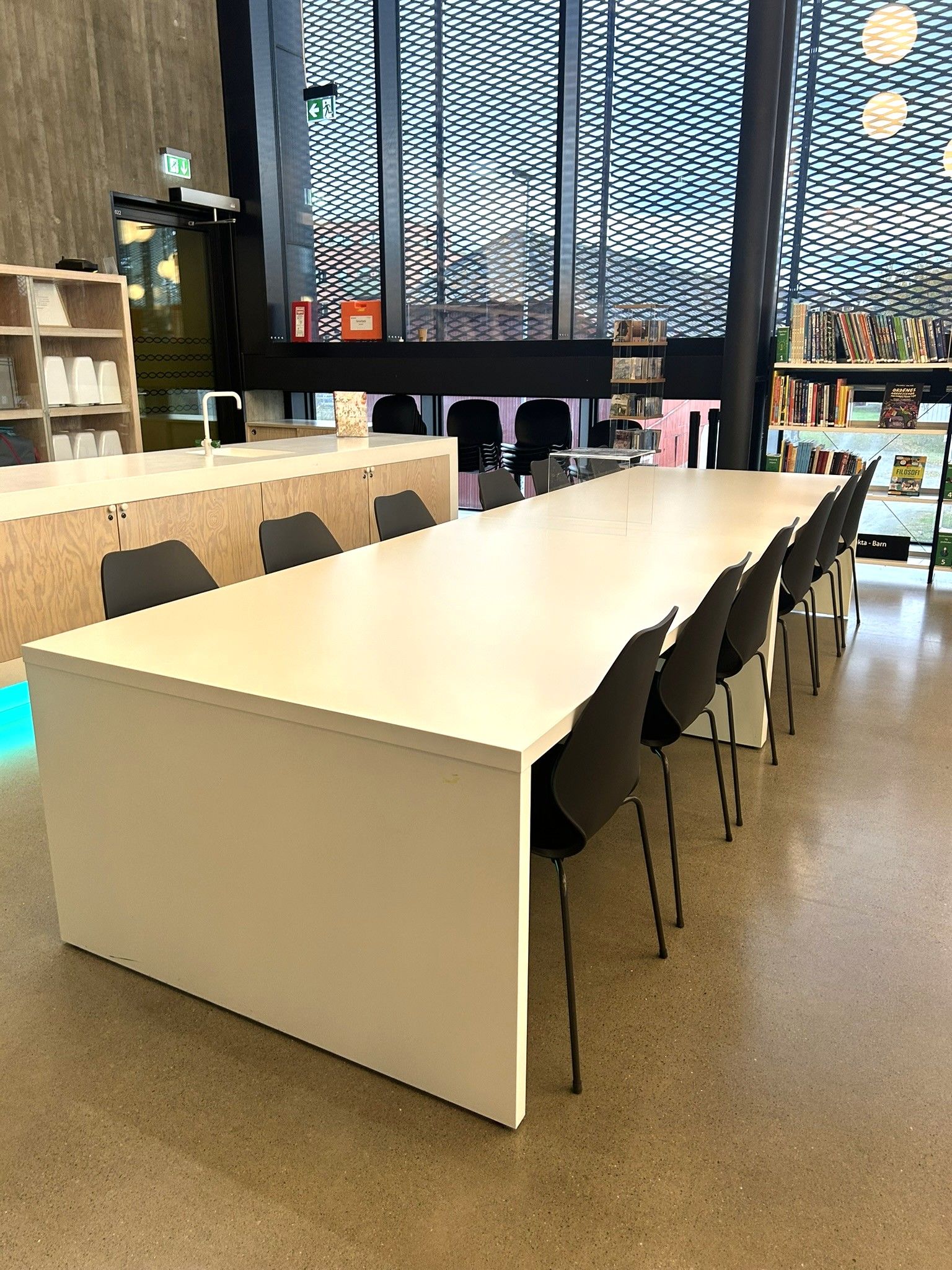 A modern library or study space with a long white table, black chairs, and large windows.