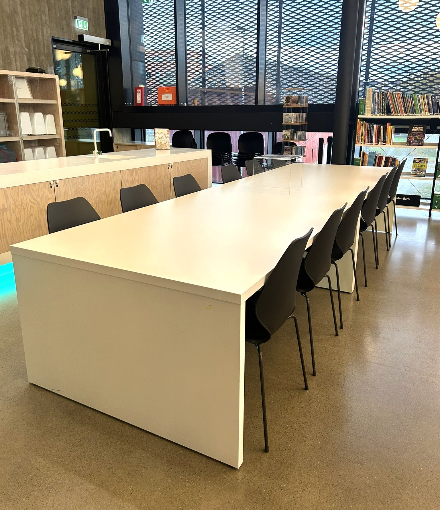 A modern library or study space with a long white table, black chairs, and large windows.