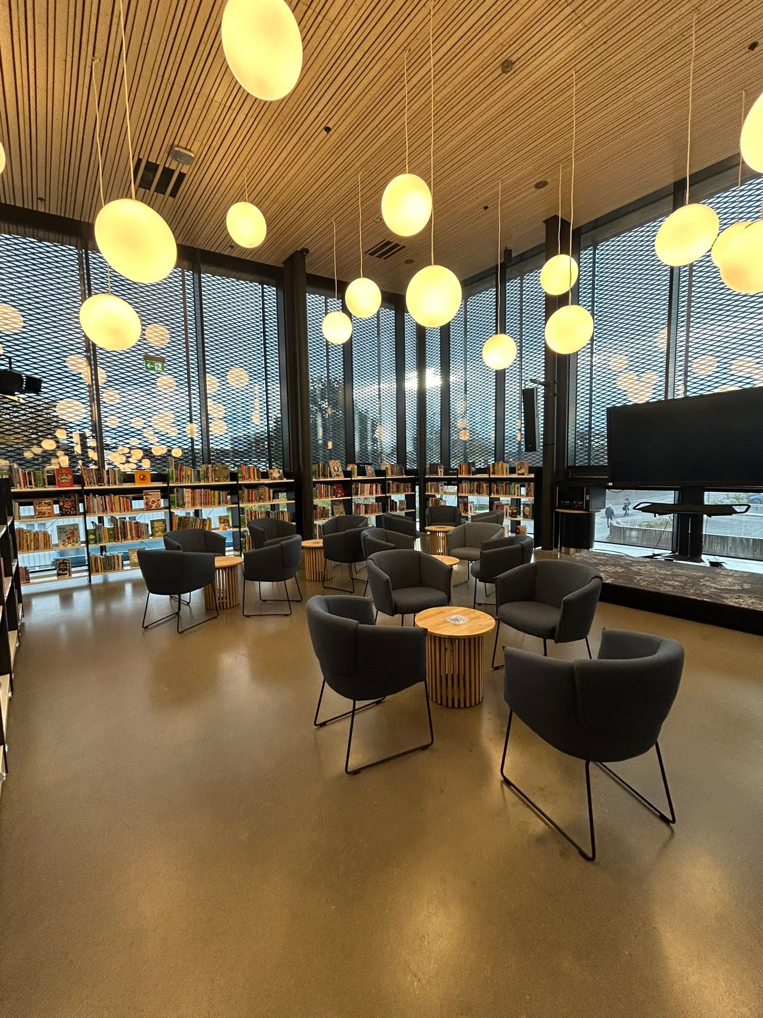Modern library lounge with dark grey armchairs, wooden tables, spherical pendant lights, tall mesh windows, and bookshelves.