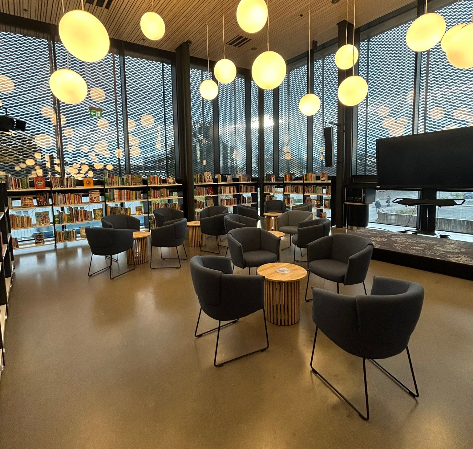 Modern library lounge with dark grey armchairs, wooden tables, spherical pendant lights, tall mesh windows, and bookshelves.
