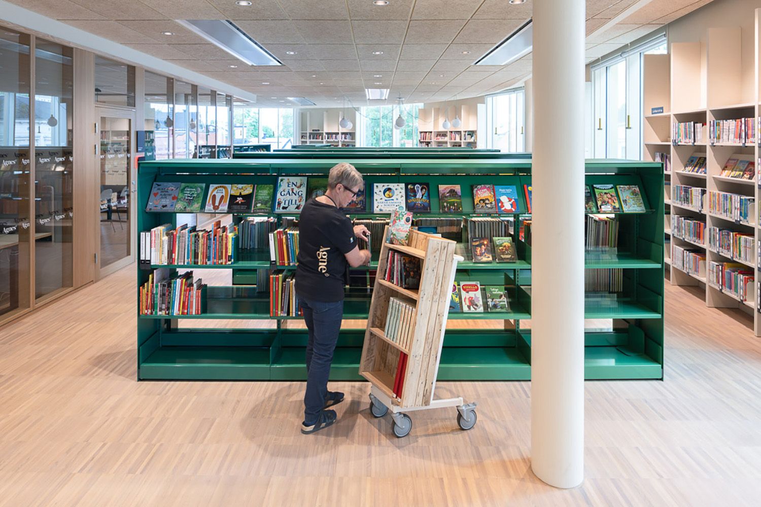 en dame står ved en bokvogn foran en grønn bokreol