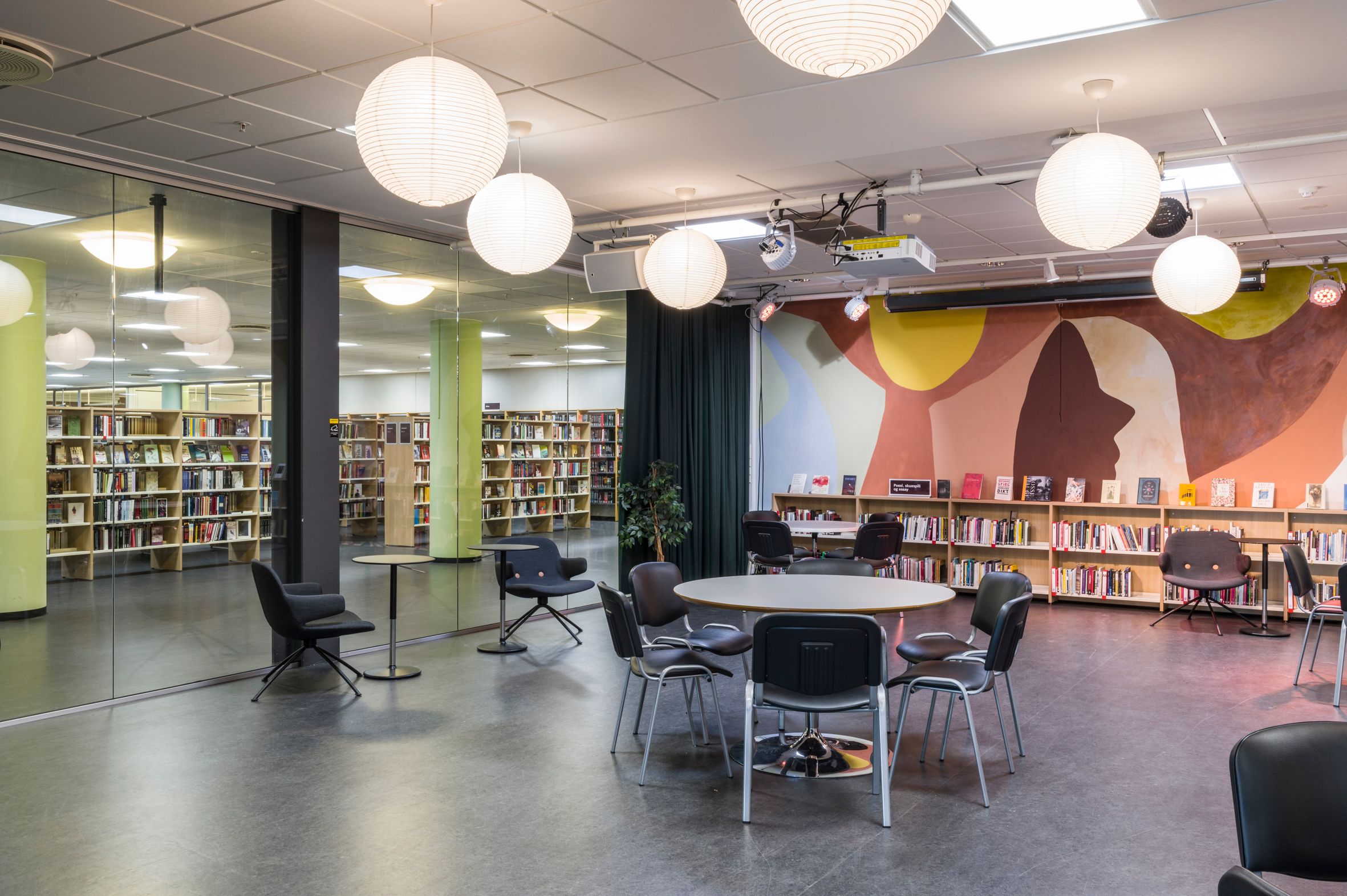 A contemporary library room with a colorful mural depicting abstract faces, rows of bookshelves, and seating areas with tables and chairs.