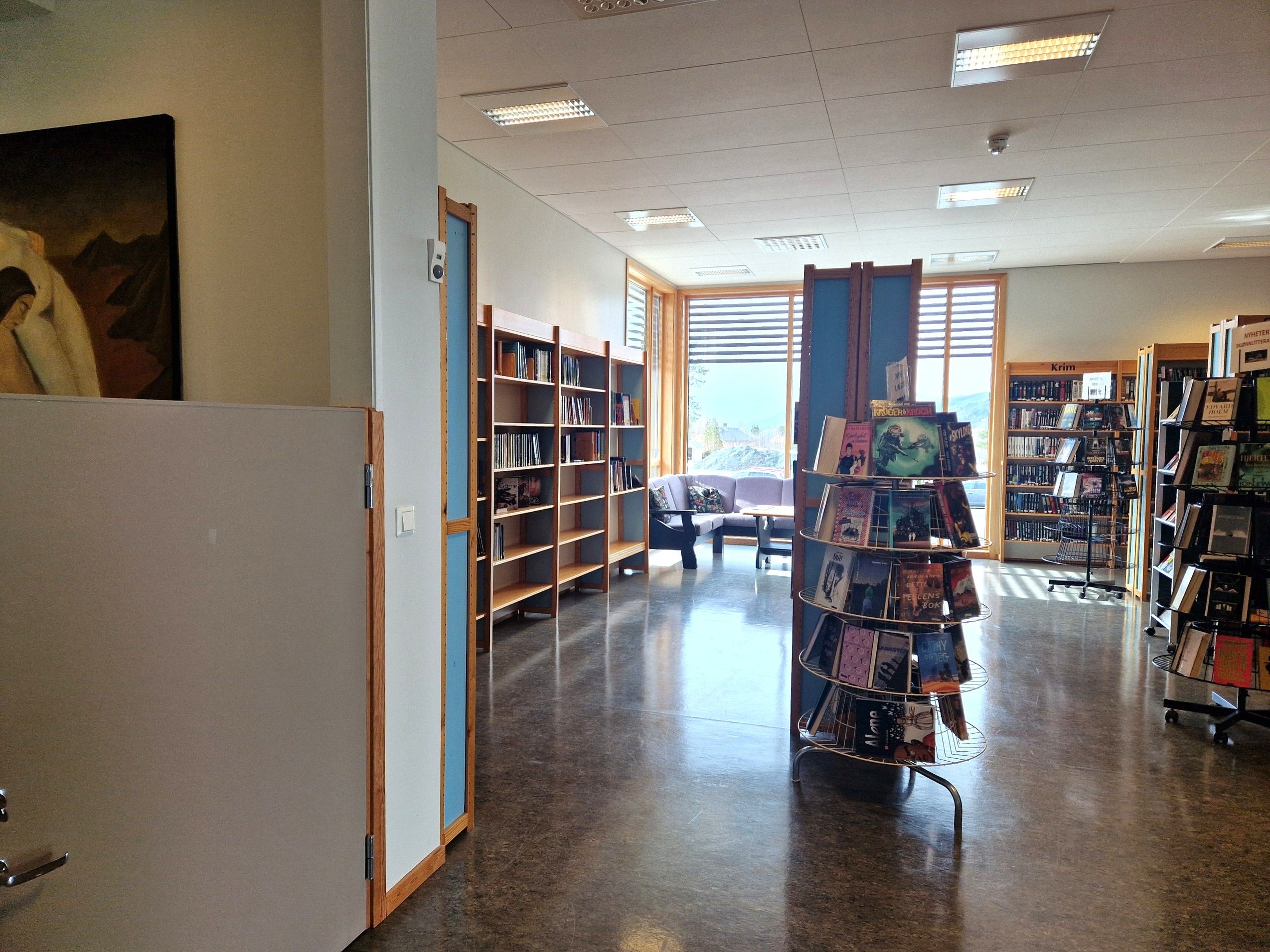 A spacious library interior with bookshelves, a rotating book display, and a lounge area by windows.