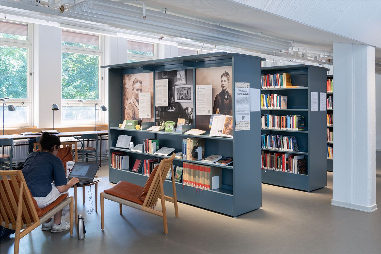 A person works on a laptop in a bright library with bookshelves and a historical exhibit.