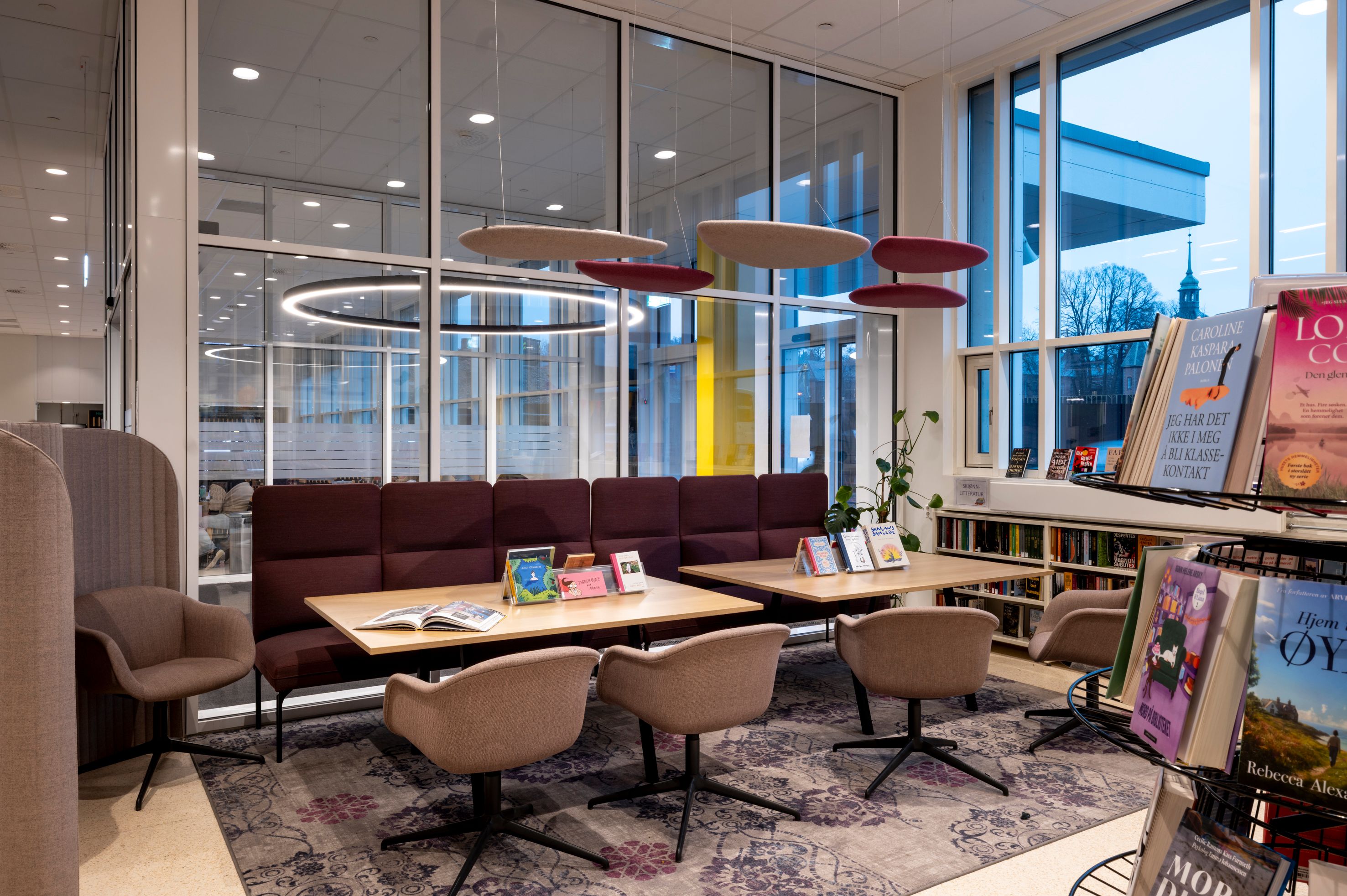 A modern library lounge with maroon booth seating, individual chairs, tables with books, and large windows.