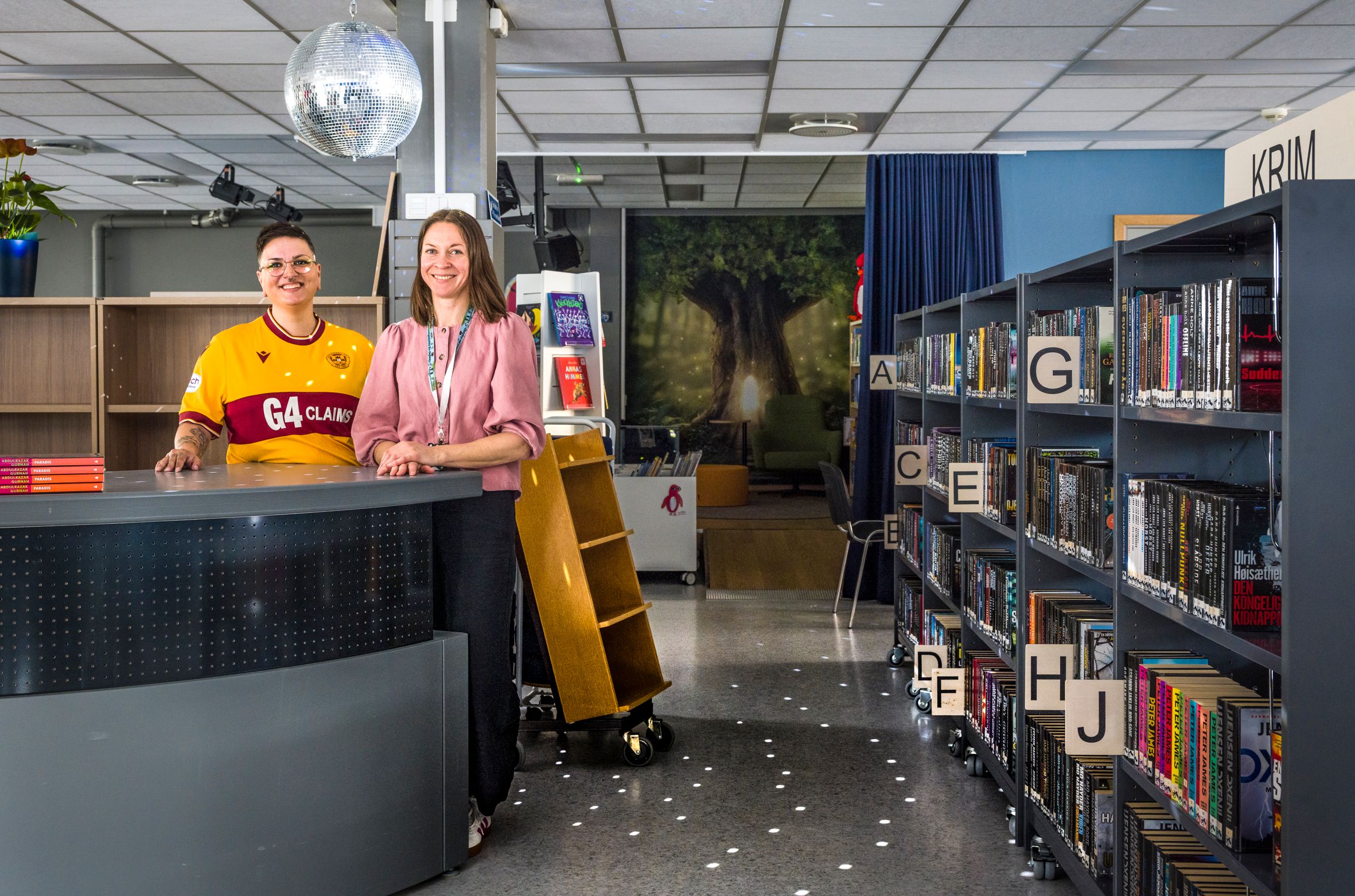 Two women smile behind a counter in a modern library with bookshelves, a tree mural, and a disco ball.