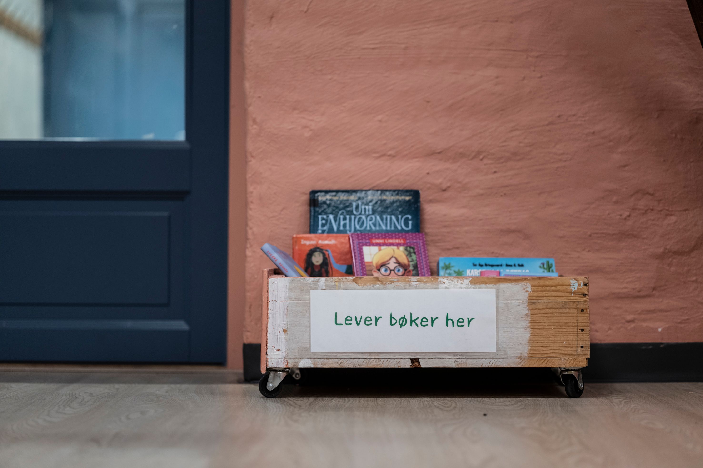A wooden box on wheels filled with books, with a sign reading "Lever bøker her" (Deliver books here).