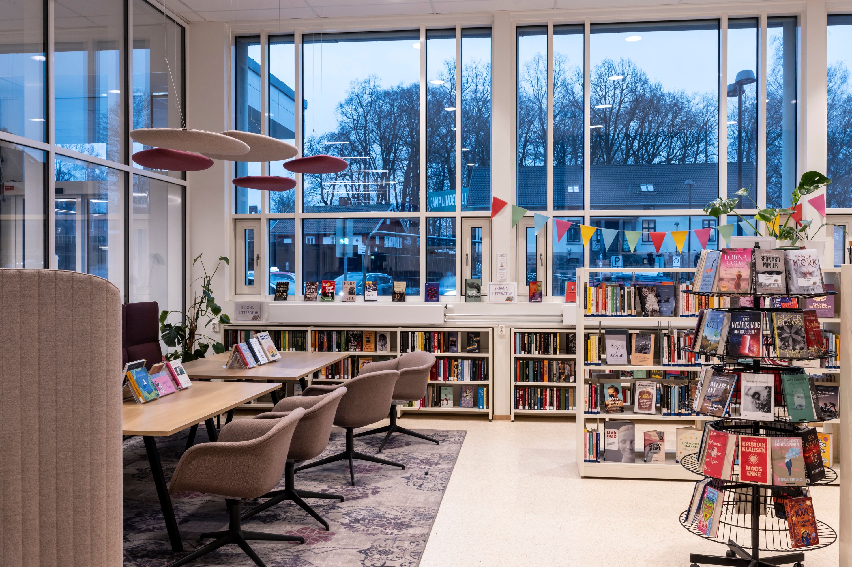 A modern library interior featuring large windows, numerous bookshelves, reading tables with chairs, a circular book display, and colorful bunting.